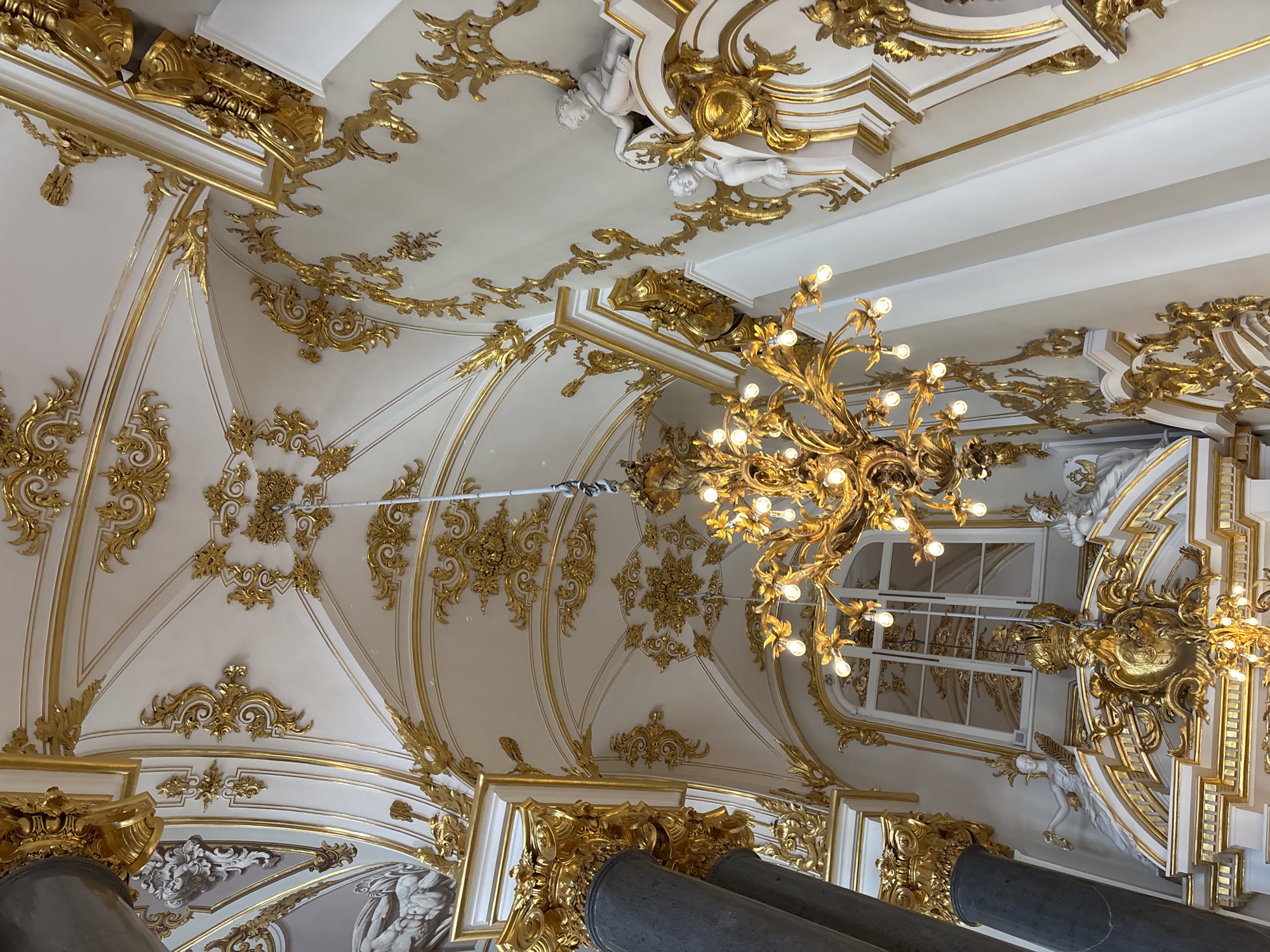 An ornate gilded chandelier hangs from a heavily decorated ceiling in a grand hall. The ceiling is adorned with intricate gold scrollwork, stucco details, and recessed lighting. A large, arched window with gold trim is visible in the background. White statues of cherubic figures are integrated into the wall and ceiling decorations. A dark, fluted column with a gilded capital is present on the left side of the image. The overall impression is one of opulence and classical architecture.