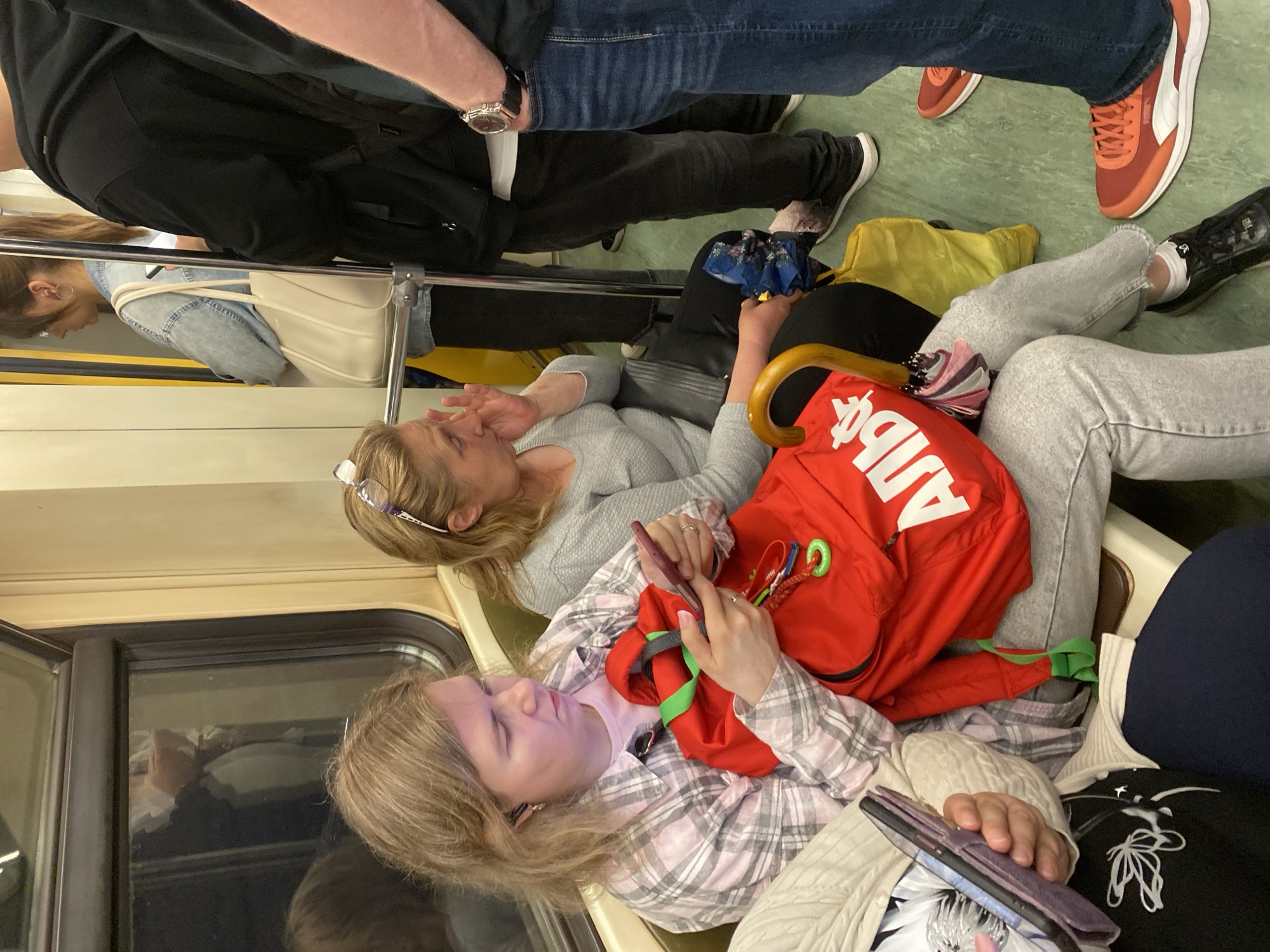 The image depicts the interior of a Moscow Metro train car during daylight. The scene is crowded, with several individuals visible. In the foreground, a woman with blonde hair, wearing a pink and grey plaid shirt, is seated and looking at her smartphone. Beside her, a red backpack with green trim is prominent. To her right, another woman with blonde hair and glasses perched on her head is seated and touching her face. Other passengers are standing and seated further back in the car, some with bags and backpacks. The train car features green seating and yellow poles.