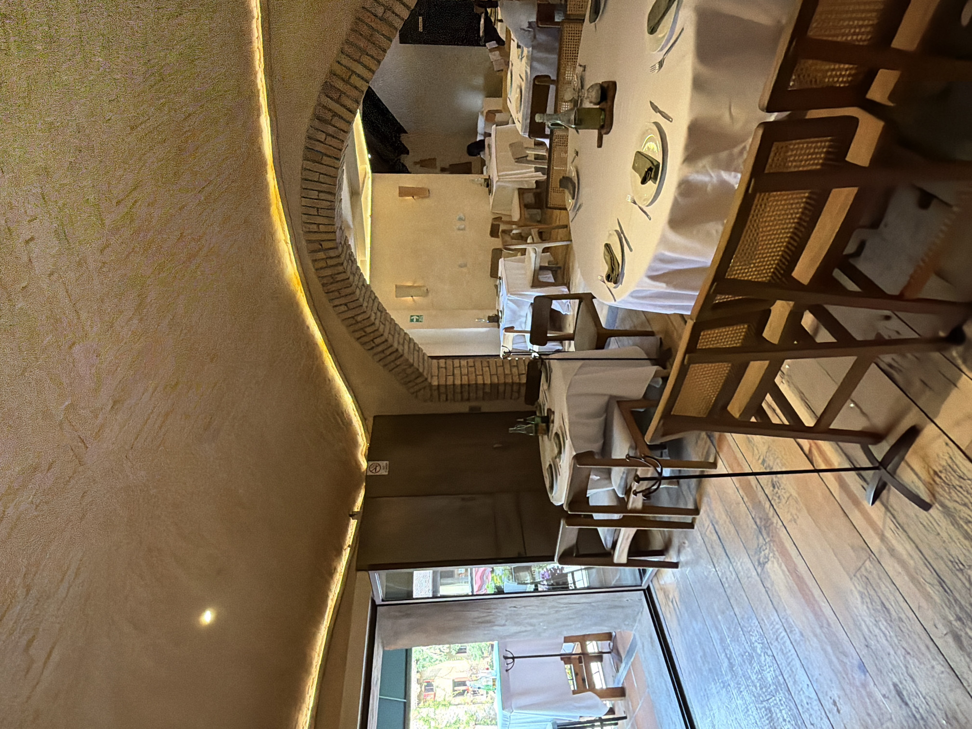 The image depicts the interior of a restaurant in Domingo Arenas, Mexico. The perspective is skewed, with the viewer looking down a long, curved dining area. The walls are finished in a textured, earthy tone, accented by an arched brick structure that serves as a doorway or transition between sections of the restaurant. The lighting is warm, possibly from recessed fixtures or ambient light sources.
Tables are set with white tablecloths and appear ready for service. The chairs are a simple, wooden design with woven seats. The flooring is wooden, likely planks. Through a doorway at the end of the dining area, there is a glimpse of an outdoor area with greenery and possibly additional seating. The restaurant seems to be well-lit and tidy. The overall atmosphere is upscale and inviting. There are no people immediately visible in the frame, but the setting suggests a dining establishment prepared for customers.