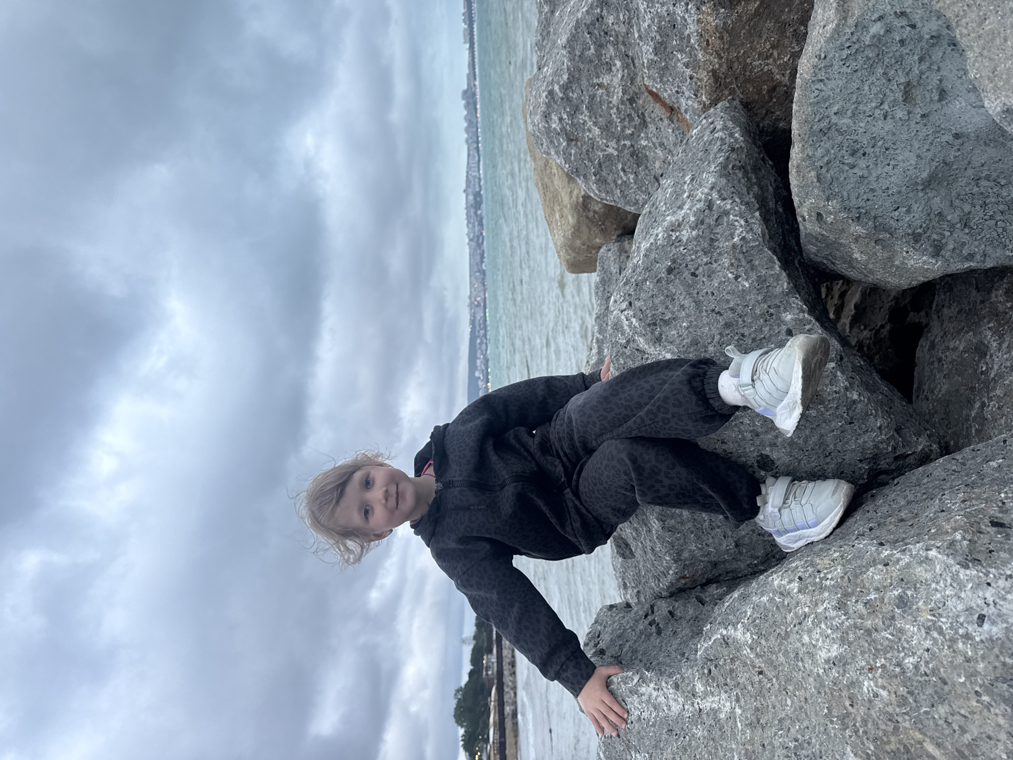A young child is sitting on a pile of large rocks with water and a city skyline in the background. The child is wearing a dark patterned tracksuit and light-colored shoes. The sky is overcast. The location is Unknown, Unknown.
