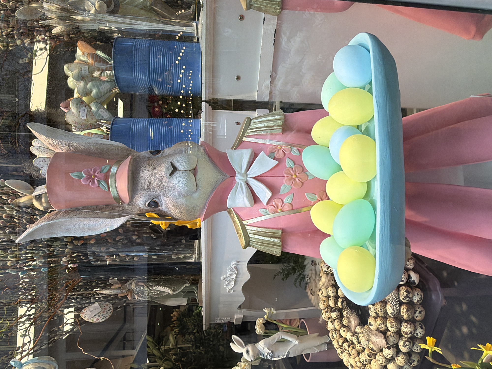 A retail window display in Luxembourg City, Luxembourg, features a large, anthropomorphic rabbit statue centrally positioned, dressed in a pink military-style uniform with gold epaulets, a white bow tie, and a pink top hat adorned with a flower. The rabbit holds a light blue, oval-shaped tray containing approximately fifteen pastel yellow and light blue decorative eggs. To the left, a smaller white rabbit statue in a suit stands beside a mound of spotted quail eggs and yellow daffodils, set against a white, ornate decorative console. To the right, two ribbed blue containers hold multiple heart-shaped decorations on sticks, some displaying price tags. The background includes bare branches with pussy willows and hanging decorative Easter eggs. Reflections of exterior elements and internal lighting, including string lights, are visible on the glass. The scene depicts an Easter-themed presentation.
