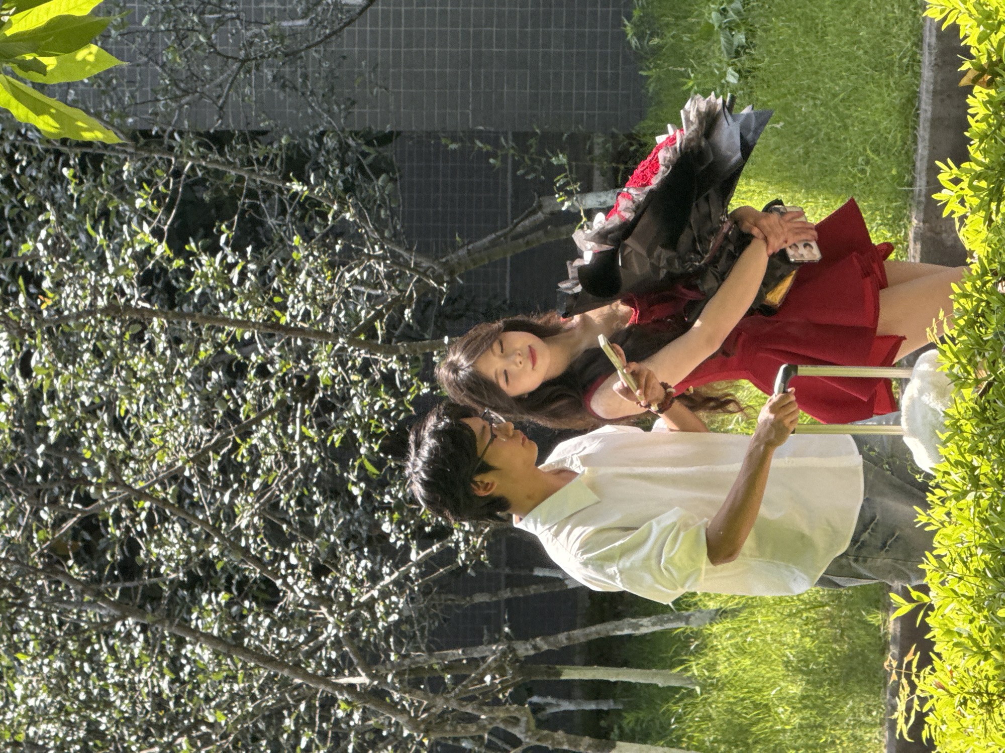 A young East Asian couple is posing for a photograph outdoors. The man is wearing glasses, a white shirt, and jeans. The woman is wearing a red dress with black lace. They are standing on a paved surface with grass and trees in the background. The location is possibly within Jiangjin, China. The man is holding a phone, presumably taking the photo. The woman is holding a phone in one hand and what appears to be a small tube or vial in the other. They are both looking at the camera.