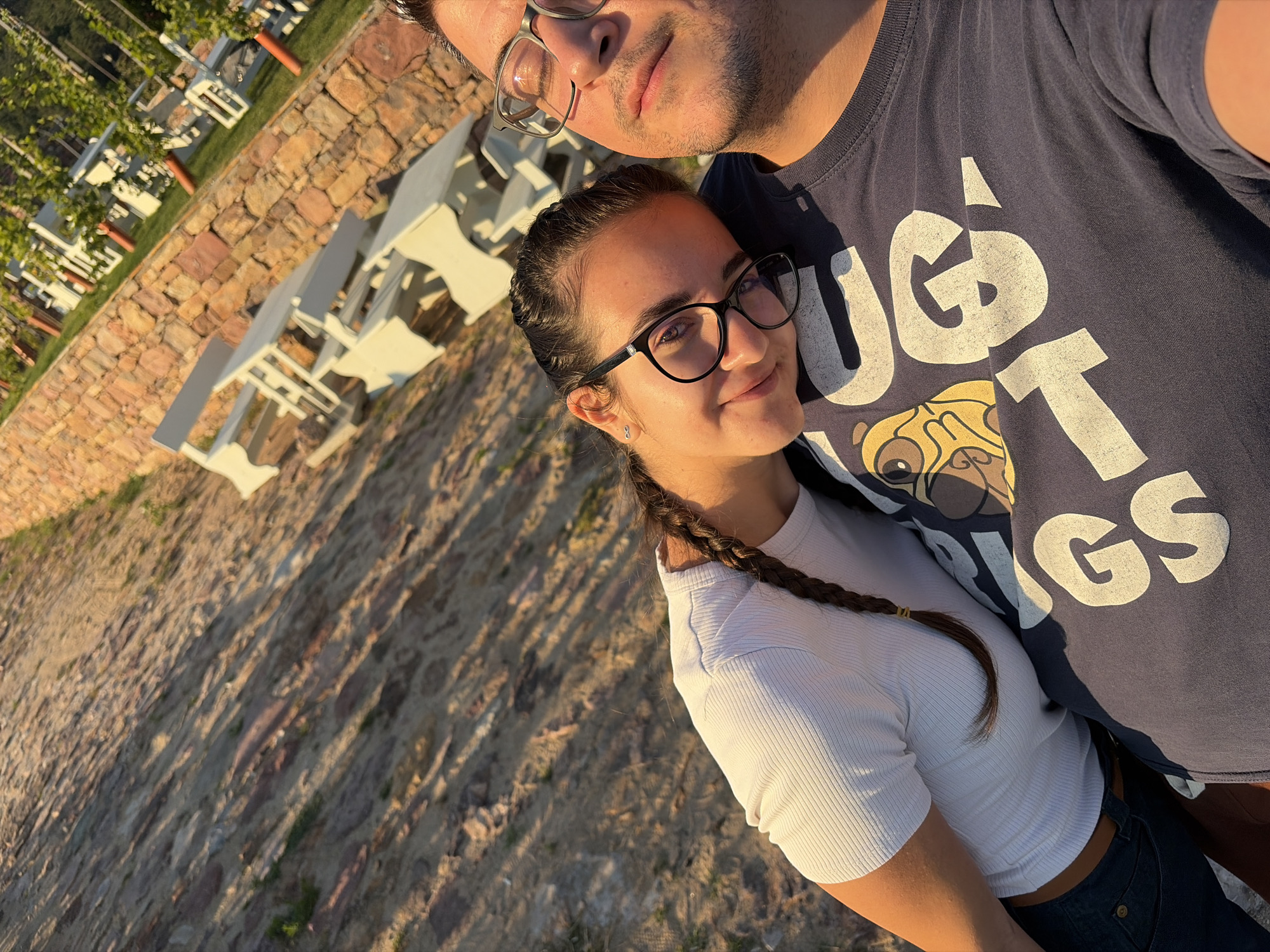 In a close-up selfie taken outdoors during golden hour, a young woman with dark hair in a braid rests her head on the shoulder of a man wearing glasses and a dark blue t-shirt with a cartoon graphic and large white lettering. The woman has on a white ribbed t-shirt and the man appears to be wearing a dark blue t-shirt. Behind them, a stone wall separates a textured, sun-drenched ground from a grassy area with white picnic tables and some greenery, possibly a vineyard, in the background. The lighting suggests a warm, late afternoon or early evening setting. The backdrop's features are consistent with an outdoor recreational area within Sofia, Bulgaria, likely a park or a winery.