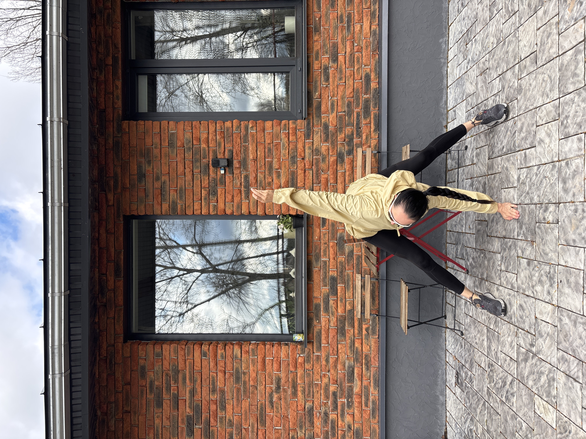 This image, taken in Chaikovsky, Russia, features a person engaged in an athletic or yoga-like pose on an outdoor paved area next to a modern brick building. The photo is rotated, presenting a dynamic, high-angle view.
The central subject is an individual, likely female, with long dark braided hair, dressed in a light yellow/khaki windbreaker jacket, black leggings, and dark athletic shoes. They are captured in a wide-legged stance with arms extended horizontally, parallel to the ground, and palms facing downwards, suggesting a balanced and focused posture.
The setting is an outdoor patio paved with rectangular grey stones. To the left, a contemporary building constructed from dark red bricks with dark grey mortar provides a backdrop. The brick wall features two large, dark-framed windows that reflect the bare branches of trees against a cloudy, overcast sky, indicating either late autumn or winter. A silver-colored downspout is visible along the left edge of the building. Partially visible behind the person are elements of outdoor furniture, including two small chairs with dark metal frames and wooden slats. To the right of the brickwork, a section of dark grey, textured wall completes the background.
The lighting is soft and diffused, characteristic of an overcast day, with no harsh shadows. The reflections in the windows are a notable detail, clearly showing the stark silhouettes of leafless trees and a cloudy sky, suggesting cool weather. No discernible text is visible in the image.