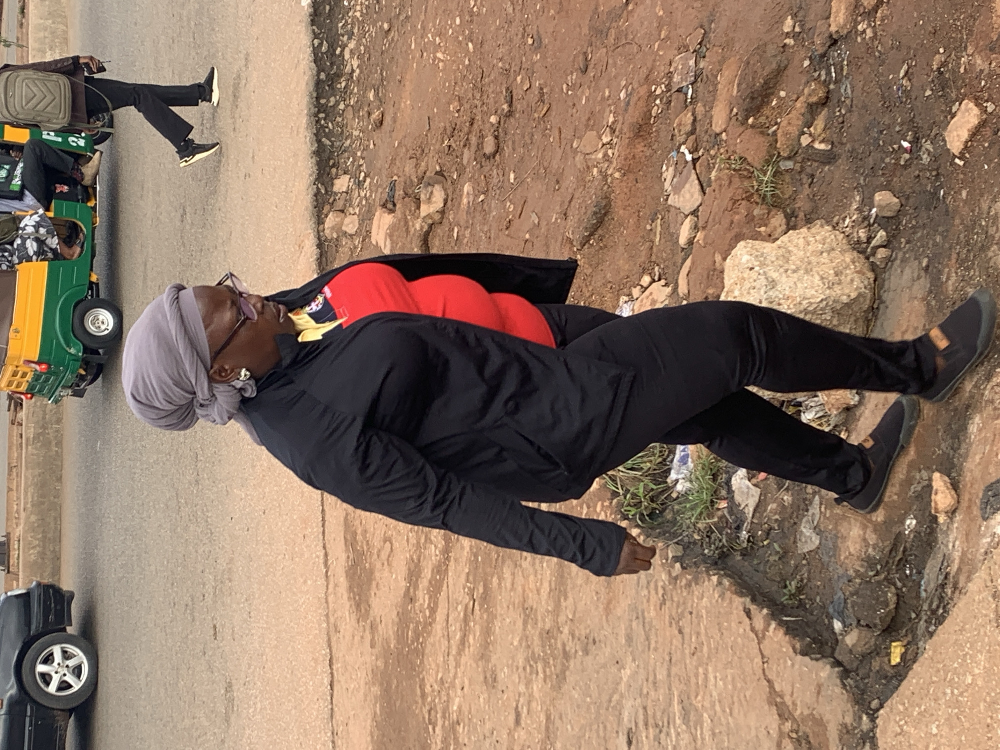 In Gura Top, Nigeria, a woman with a grey headscarf and glasses walks on a dirt path beside a road. She wears a black jacket over a red shirt and black pants. A black car is visible on the road to her left, and a green and yellow auto-rickshaw with passengers is on the road to her right, with a man in a brown jacket and a backpack walking behind it. The environment is unpaved and appears to be a street with debris and rocks.