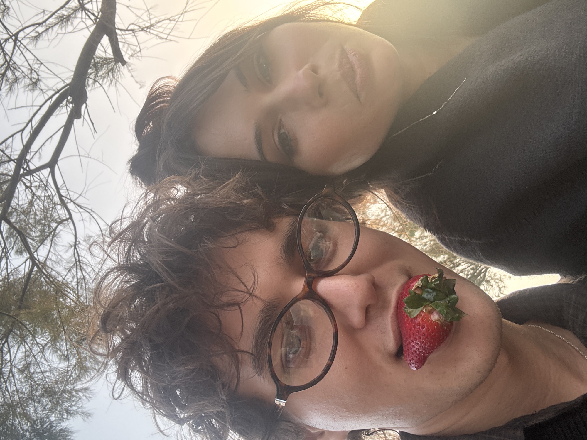 A close-up, low-angle selfie shows two individuals: a woman and a man. The man is in the foreground, sporting round glasses, curly hair, and holding a strawberry in his mouth. The woman is positioned slightly behind and above him. The background reveals a hazy, bright sky partially obscured by tree branches. The location appears to be outdoors, potentially in Tel Aviv, Israel, though no specific landmarks are visible in the frame to confirm it.