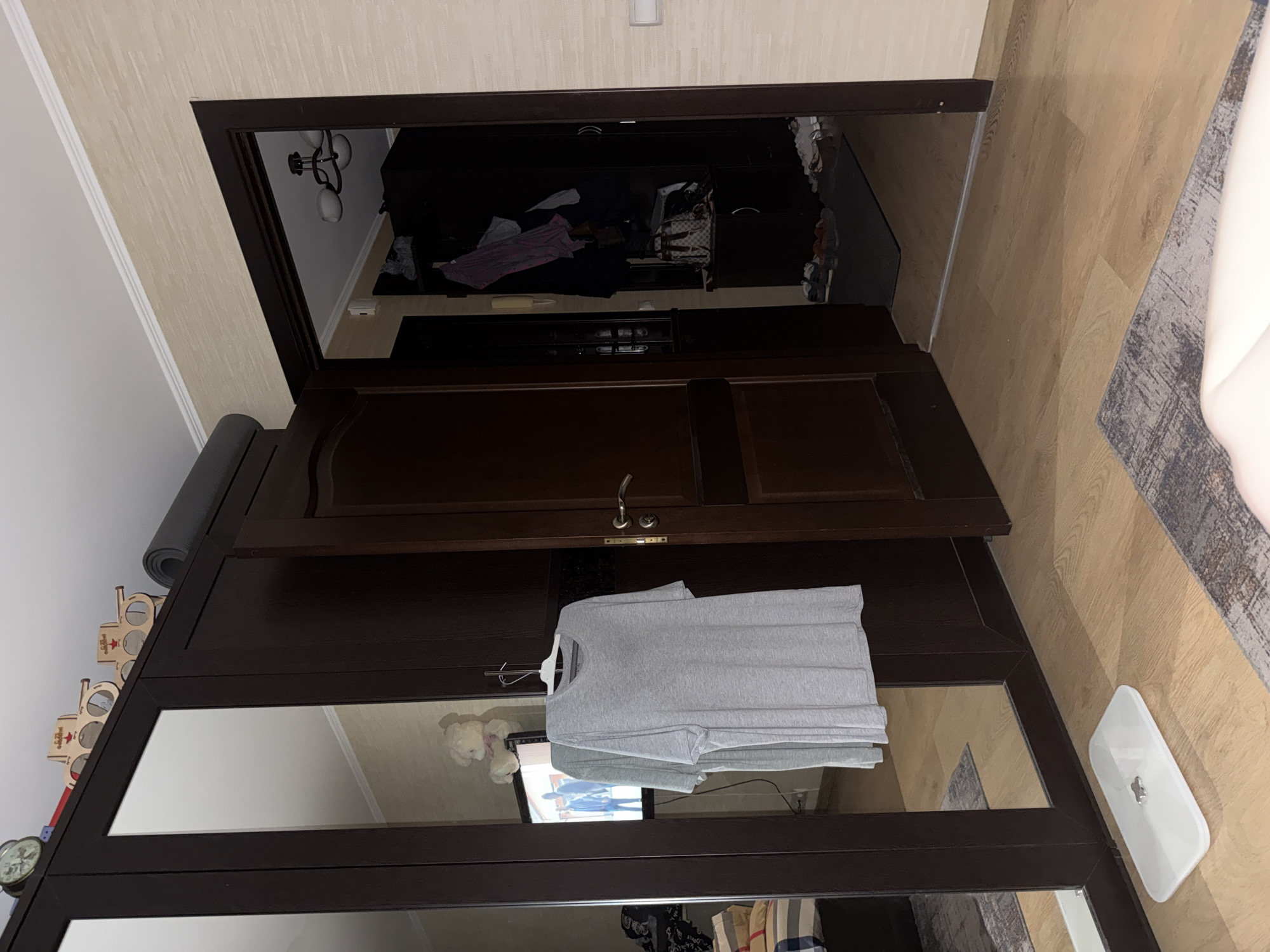 The image shows an interior scene, likely a bedroom or dressing area, with a large dark wood wardrobe that has mirrored doors. A gray t-shirt is hanging on a hanger from a hook on the wardrobe door. To the right, an open doorway leads to another room. Inside the doorway, there is a dark-colored closet filled with clothes and bags, and shoes are visible on the floor. In the foreground, a white digital scale is on the wooden floor, and a rug is partially visible. The wall to the right of the doorway is textured and light-colored. The ceiling has white trim. On top of the wardrobe, a black yoga mat is rolled up, and two wooden decorative objects resembling tanks are placed next to it. A vintage alarm clock is visible on the top left corner of the wardrobe. A television screen is visible through the mirrored door of the wardrobe, reflecting a room with what appears to be a teddy bear and framed pictures. The overall lighting suggests it is nighttime or the room is dimly lit.
