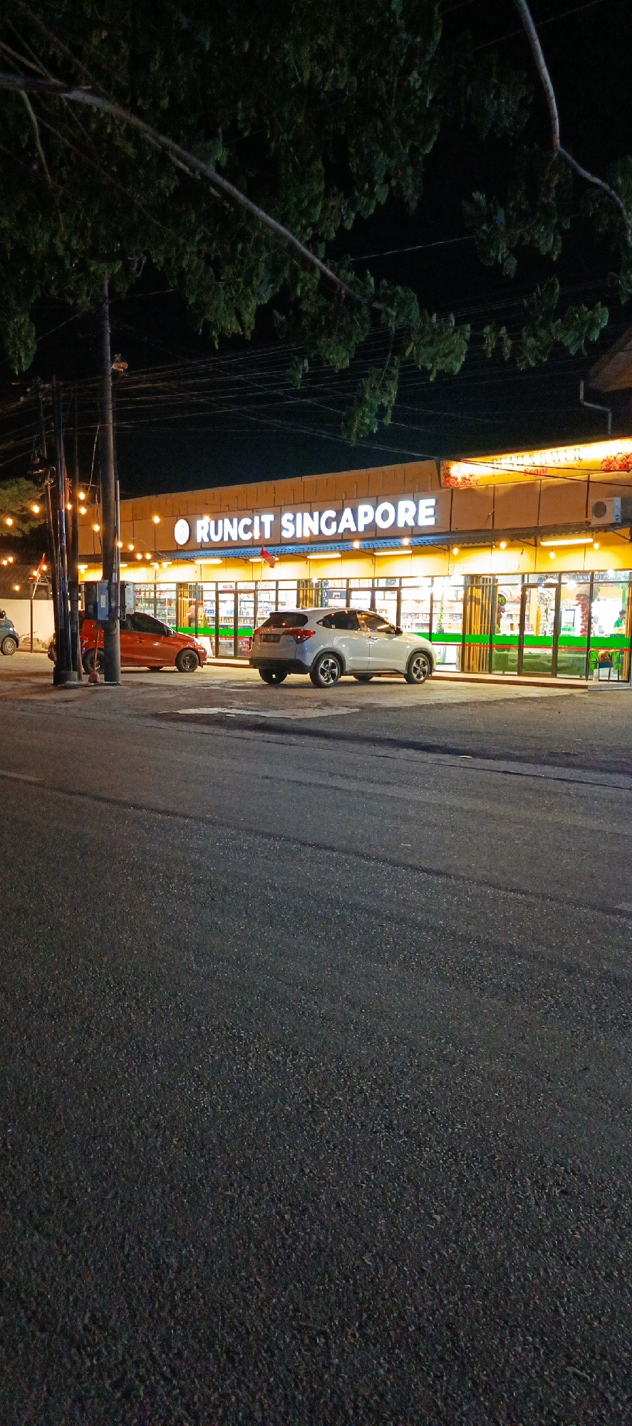 A night-time exterior shot captures the facade of a well-lit convenience store, identified by the prominent white lettering "RUNCIT SINGAPORE" above its entrance. The store appears to be located on a street in Banda Aceh, Indonesia, as indicated by a sign within the store's window that reads "TOKO MINUMAN" and "GROSIR" which translates to "drink shop" and "wholesale" respectively, and a red flag that is consistent with the national flag of Indonesia. Two cars are parked in front of the store: a red sedan on the left and a silver SUV on the right. The store's interior is visible through large glass windows and doors, revealing shelves stocked with products and overhead lighting. Several strings of decorative lights are strung along the front of the building, casting a warm glow. The street in the foreground is paved asphalt, and a tree with dark green foliage partially obscures the top of the image. Electrical poles and wires are visible on the left side of the frame. The overall scene depicts a typical street-level commercial establishment operating at night.