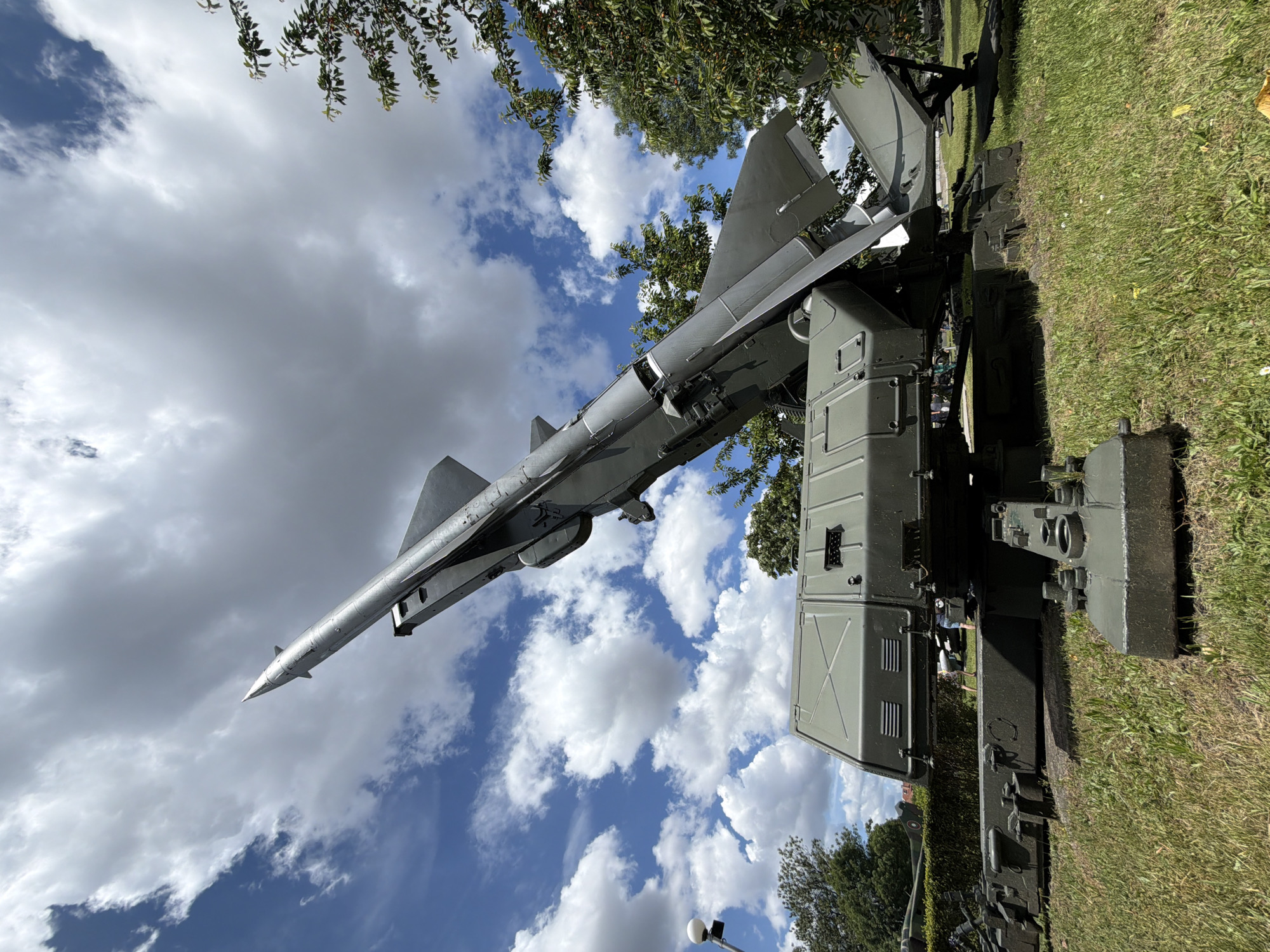 The image displays a military missile launcher in Sofia, Bulgaria, positioned outdoors on a grassy area. The launcher, painted in a drab olive green, is the central subject, with its missile angled upwards towards a dramatic cloudy sky. The missile itself is a sleek, silver projectile, contrasting with the darker green of the launcher's base and missile housing. The launcher is situated on a complex base with visible mechanical components and appears to be a static display, possibly in a museum or memorial site. In the background, trees and a lamppost are visible, along with a glimpse of a building and possibly other military vehicles further in the distance. The ground is covered in green grass with some scattered yellow wildflowers. The sky is a mix of bright blue and large, white cumulus clouds, suggesting a fair weather day. A few people are present in the background, appearing as small figures and not actively interacting with the main subject. The overall impression is one of a historical military artifact on display in a public park or exhibition area within the city of Sofia.