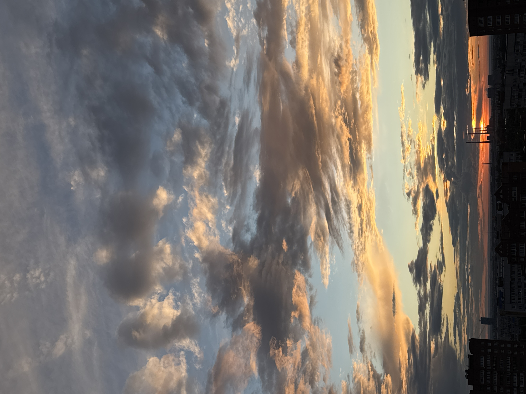 An urban landscape unfolds beneath a dramatic sky filled with scattered clouds, illuminated by the warm hues of a sunset. In the foreground, the silhouettes of apartment buildings are visible, their dark forms contrasting with the vibrant sky. The middle ground features industrial structures, including tall smokestacks, hinting at a city environment. The horizon is marked by a faint line of distant buildings and possibly hills. The sky is a canvas of grays, blues, and oranges, with clouds streaked with golden light. The overall impression is one of a city at dusk, with the natural beauty of the sunset over industrial elements.