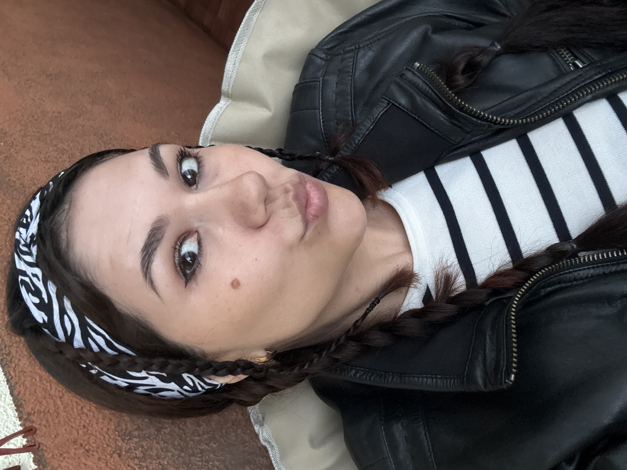 A close-up, overhead shot shows a young woman with dark hair in braids and a zebra-print bandana. She is wearing a black leather jacket over a white and black striped shirt. Her lips are pursed, and her eyes are wide open, looking directly at the camera. A small brown mole is visible on her right cheek. The background consists of a textured brown surface and a neutral-colored fabric. The lighting appears to be natural, suggesting it could be daytime. The woman's expression is somewhat playful or curious. There is no visible text in the image.
