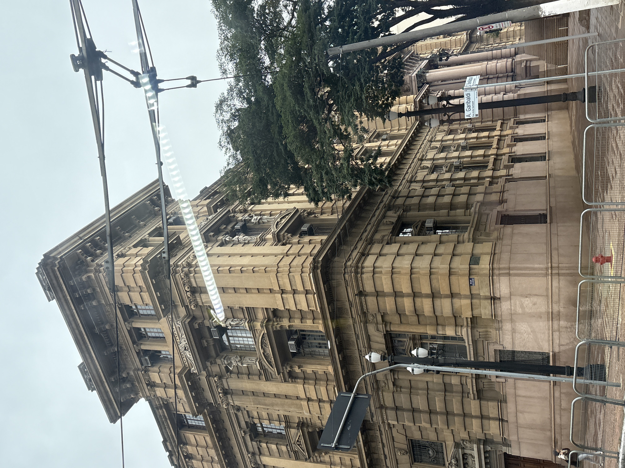 The image shows a section of a grand, historic building in the city of São Paulo, Brazil. The building is made of weathered, tan stone with intricate architectural details, including columns, moldings, and sculptures adorning its facade. It features numerous windows with dark frames, some of which have air conditioning units installed. In the foreground, there is a street scene with a lamppost, a traffic sign, and metal barriers. A person is visible in the lower left corner, walking away from the camera. Behind the building, a large tree with dense green foliage is visible. Overhead, there are power lines and equipment, likely for a public transportation system like a tram or trolleybus. Street signs indicate "RUA AV GARIBALDI" and "RUA ANTÔNIO GARIBALDI", suggesting the location is near Garibaldi Avenue. The sky is overcast, creating a subdued lighting condition. A red fire hydrant is visible near the right edge of the image.