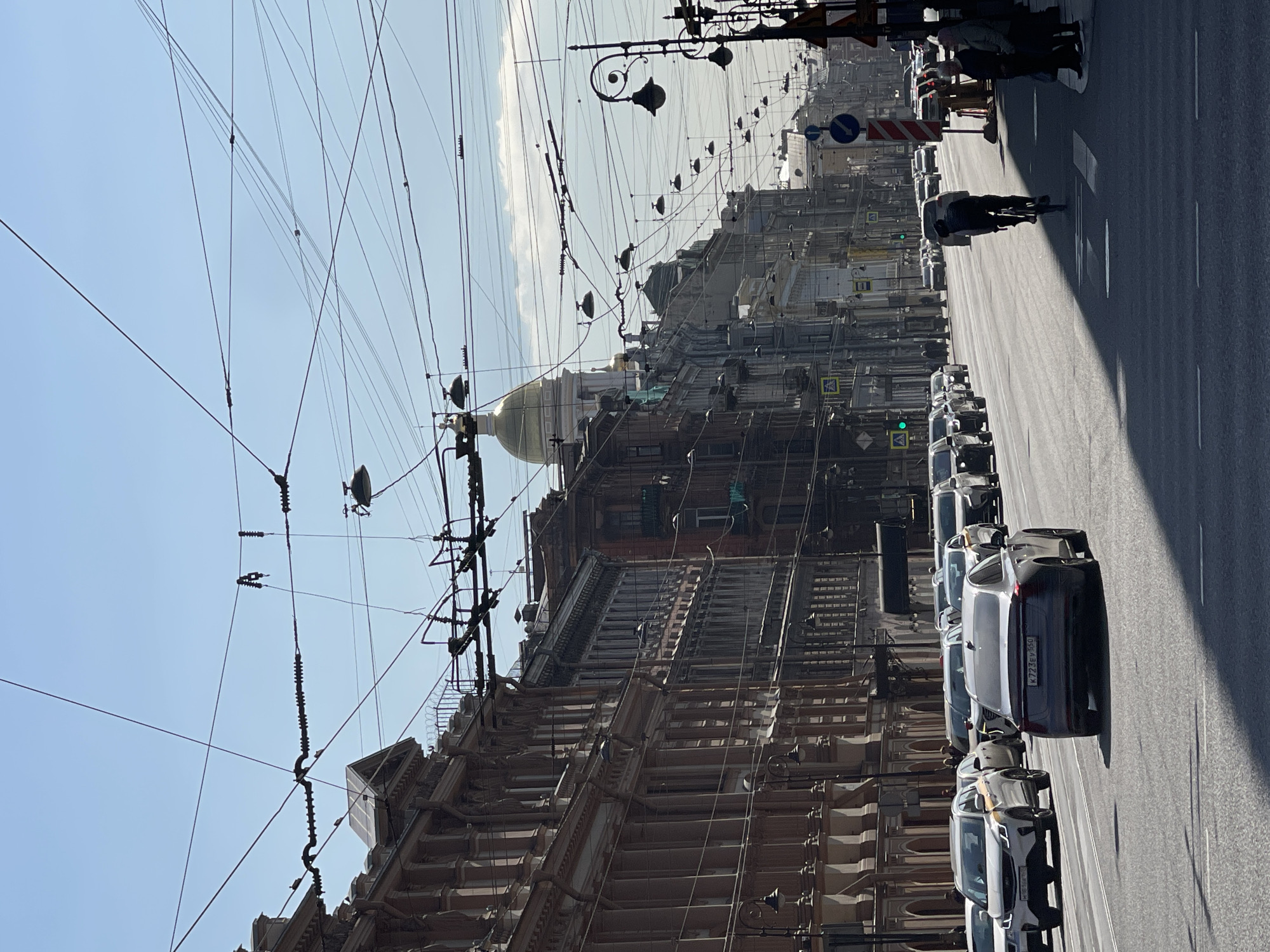 The image captures a street scene in Saint Petersburg, Russia. A wide avenue stretches into the distance, lined with buildings of ornate architectural design. Prominently visible is the dome of Saint Isaac's Cathedral. An array of overhead power lines crisscrosses the sky above the street. Cars are parked along the side of the road, and pedestrians can be seen walking and cycling on the avenue. The day appears bright and sunny, with a clear sky visible.