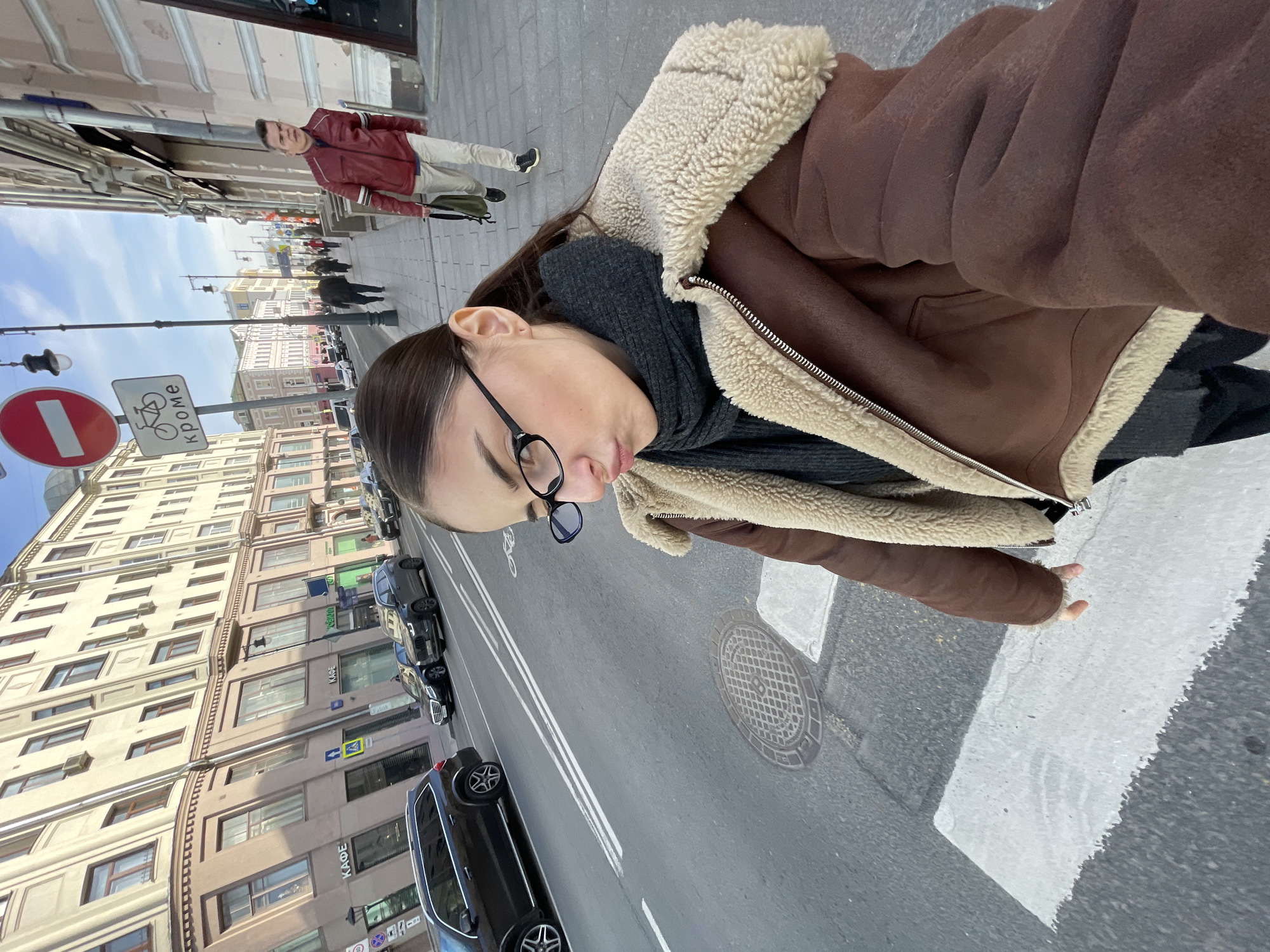 An upward-facing, close-up photograph captures a person on a street in Moscow, Russia, with an urban background. The primary subject, seen from the chest up, is a young person with dark hair, wearing glasses, a dark grey scarf, and a brown jacket with a prominent beige shearling lining. Their gaze is directed downwards, suggesting they are looking at the camera of the device used to take the selfie.
In the background, a bustling city street unfolds. To the left, several multi-story buildings with numerous windows line the street. Many cars, predominantly dark-colored, are parked or driving on the asphalt road, which features clear double white lines and a circular manhole cover. A white-striped pedestrian crossing is visible in the foreground.
Several notable details are present:
* A red circular sign with a white horizontal bar, typically indicating "No Entry" or "Do Not Enter."
* Adjacent to it, a rectangular sign features a bicycle symbol and the Russian word "КРОМЕ" (KROME), meaning "except" or "besides," implying "No Entry Except Bicycles."
* Further down the street, buildings display text, including "КАФЕ" (CAFE) on a storefront.
* Another pedestrian crossing sign, a yellow diamond shape with a black pedestrian symbol, is visible further down the street.
* Other pedestrians are present, including a man in a red jacket and light trousers walking away from the camera in the mid-ground.
* The sky appears clear and bright, indicating daytime. One car appears to have "ЯНДЕКС GO" (YANDEX GO) partially visible, suggesting a ride-hailing service.