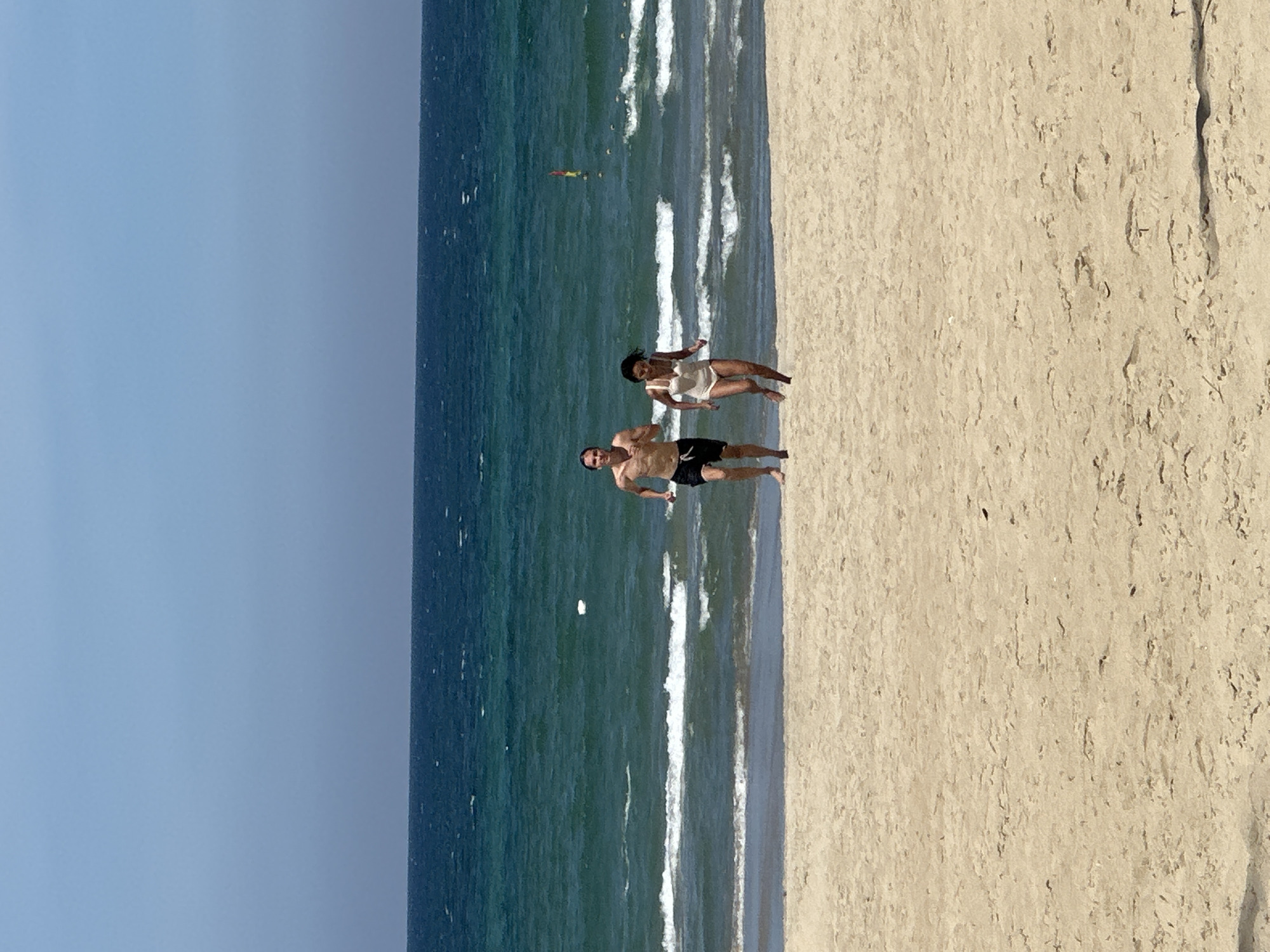 Two individuals are running towards the camera on a sandy beach, with the turquoise ocean and a clear blue sky as the backdrop. The person on the left is a male wearing black swim shorts, and the person on the right is a female in a white swimsuit. Waves are breaking on the shore behind them. The scene is consistent with a view of My Khe Beach in Da Nang, Vietnam, a popular destination known for its long stretches of sand and vibrant blue waters.