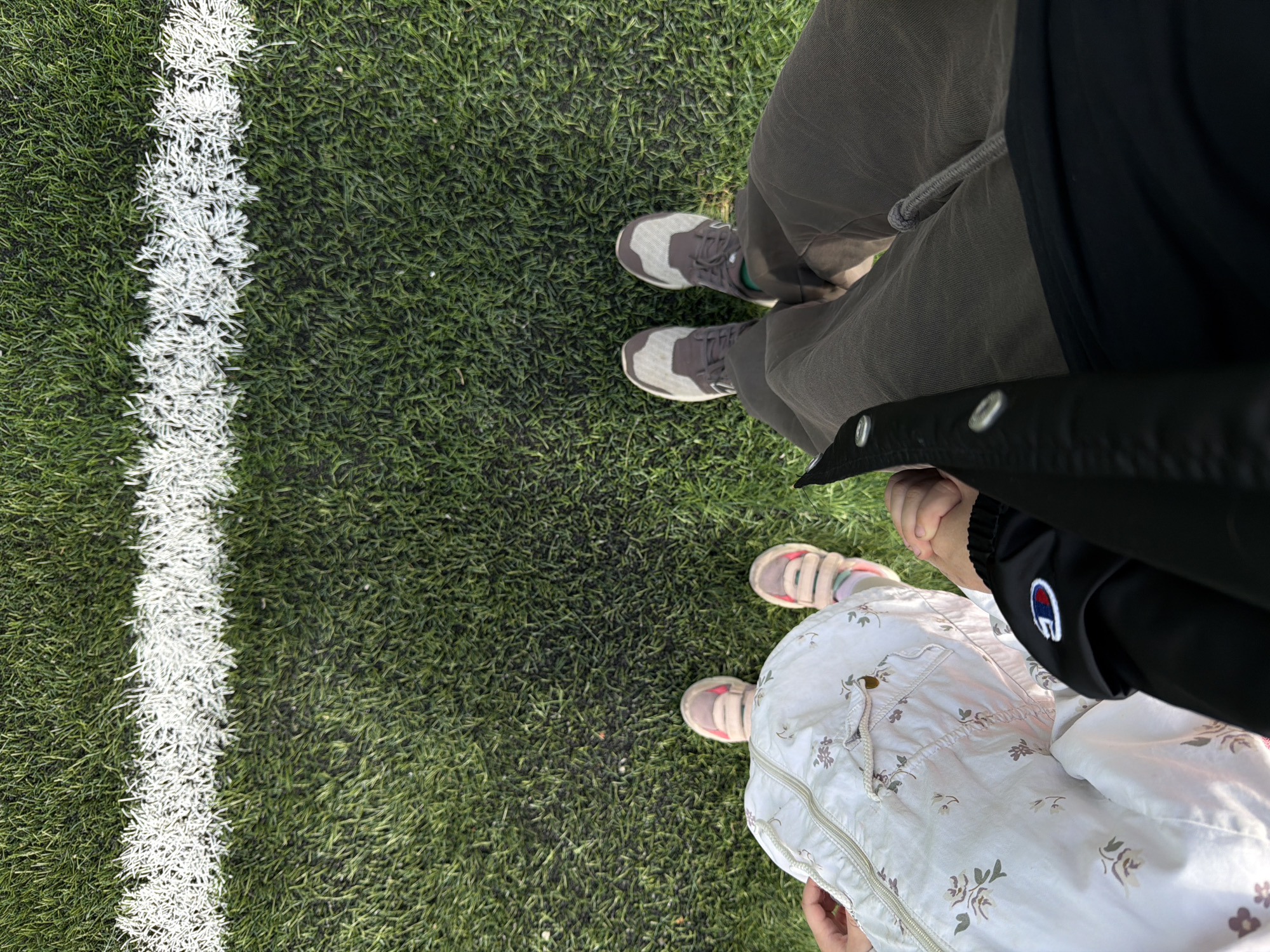 Two individuals, one adult and one child, are standing on artificial turf, likely a sports field. The adult is wearing dark pants and sneakers, while the child is wearing a light-colored jacket with a floral pattern and pink sandals. They are holding hands, suggesting a familial relationship. A white line, likely marking a boundary or play area, is visible across the top portion of the image. The background consists of green turf, indicating an outdoor setting. There are no specific indicators in the image to pinpoint the location to Bykovo, Russia.