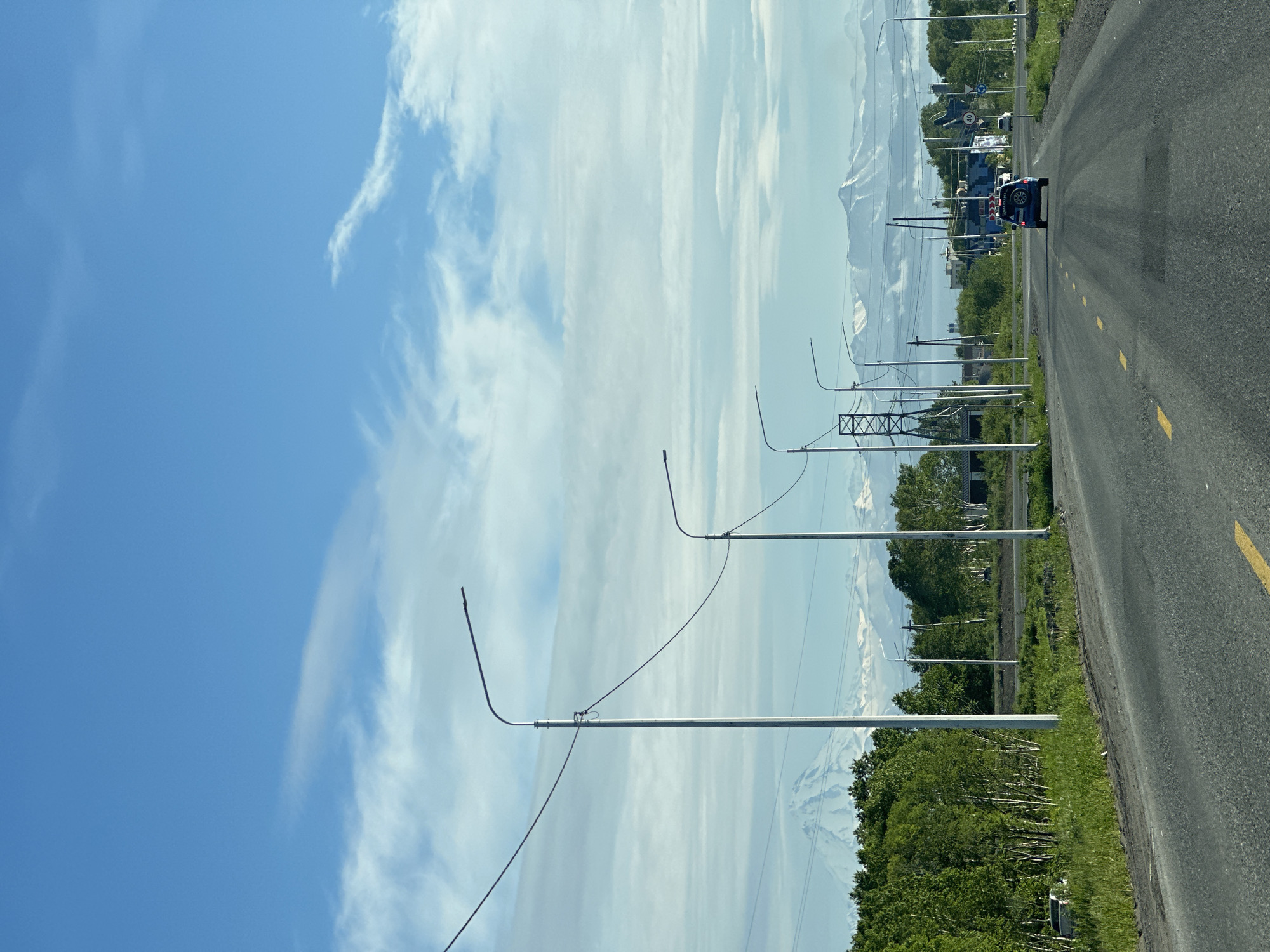 The image depicts a road in the city of Svetly, Russia, under a partly cloudy blue sky. The road, which is asphalted and has yellow lane markings, stretches into the distance. Several streetlights line the right side of the road, with power lines running between them. On the left, a dense line of green trees forms a backdrop, and beyond the trees, snow-capped mountains are visible. A blue SUV is driving away from the camera on the road. In the middle ground, there are various structures, including what appear to be buildings and power transmission towers. The overall scene suggests a semi-rural or suburban environment, possibly on the outskirts of the city, with a dramatic mountainous landscape in the background.