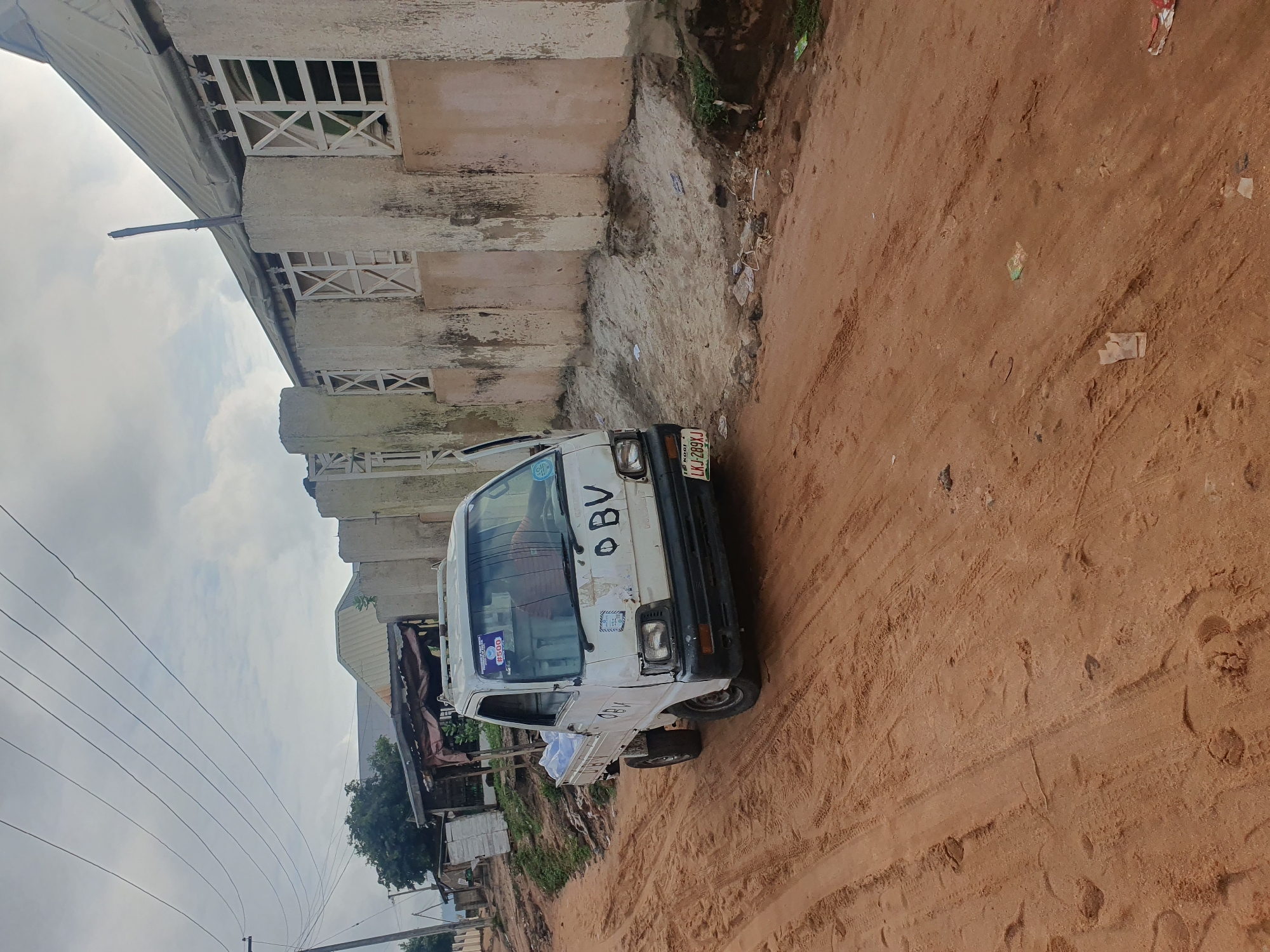 The image depicts a white mini-truck, labeled "OBV" on its windshield, parked on a reddish-brown, dirt road in what appears to be a residential or commercial area of Lokoja, Nigeria. The vehicle's license plate reads "LKJ-289KJ" and is a Kogi State plate. The truck appears to be used for transporting goods and is parked adjacent to a weathered, multi-story building with barred windows. There are utility poles and lines in the background, along with trees and other buildings. The road is covered in tracks, indicating frequent traffic. The sky is overcast, suggesting a cloudy day. The building has a white exterior with signs of wear. Trash is visible alongside the building. The overall setting suggests a lower-income neighborhood or commercial district within Lokoja.