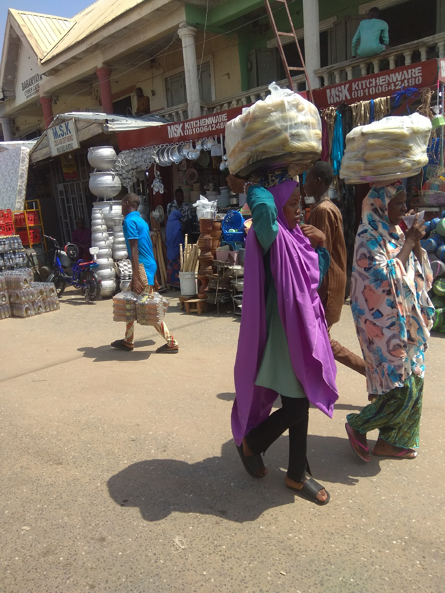 In Jalingo, Nigeria, an outdoor market scene unfolds. The sun casts shadows on the asphalted ground. A man in a blue t-shirt and patterned trousers walks to the left, carrying a large bundle of bottled drinks wrapped in plastic. To his right, a woman in a purple hijab and green garment walks forward, balancing a large tray of bagged goods on her head. Behind her, another woman in a colorful headscarf and patterned skirt walks in the same direction, holding something to her face. In the background, market stalls display various wares, including kitchenware and stacked containers. A ladder leans against a building on the right. A motorcycle is parked near stacks of large metal pots. The overall impression is of a busy commercial area on a bright day.