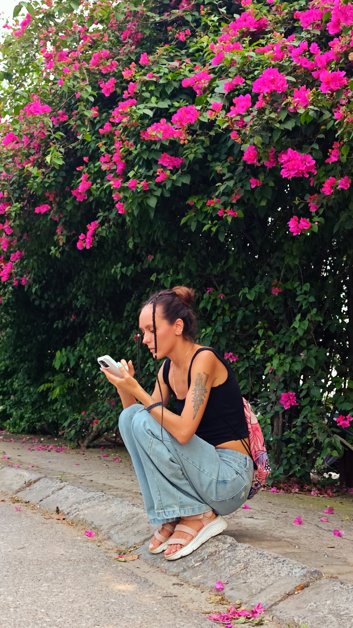 A young woman is crouching down next to a vibrant hedge of bright pink bougainvillea flowers. She has her hair pulled up in a bun with some braids around her face. She is wearing a black tank top, light blue wide-leg jeans, and light pink sandals. On her left arm, she has a tattoo. A colorful backpack is slung over her left shoulder. She is holding a white smartphone in her hands and appears to be looking at it. The ground in front of her is a paved sidewalk with a curb, and scattered pink flower petals are visible on the ground and sidewalk. The scene suggests a warm, bright day, possibly in the late morning or afternoon, given the lighting. The bougainvillea flowers indicate a tropical or subtropical climate, consistent with the provided location context of Hanoi, Vietnam. The woman's posture and focus on her phone suggest a moment of personal engagement with technology amidst a beautiful natural backdrop. There is no visible text in the image.