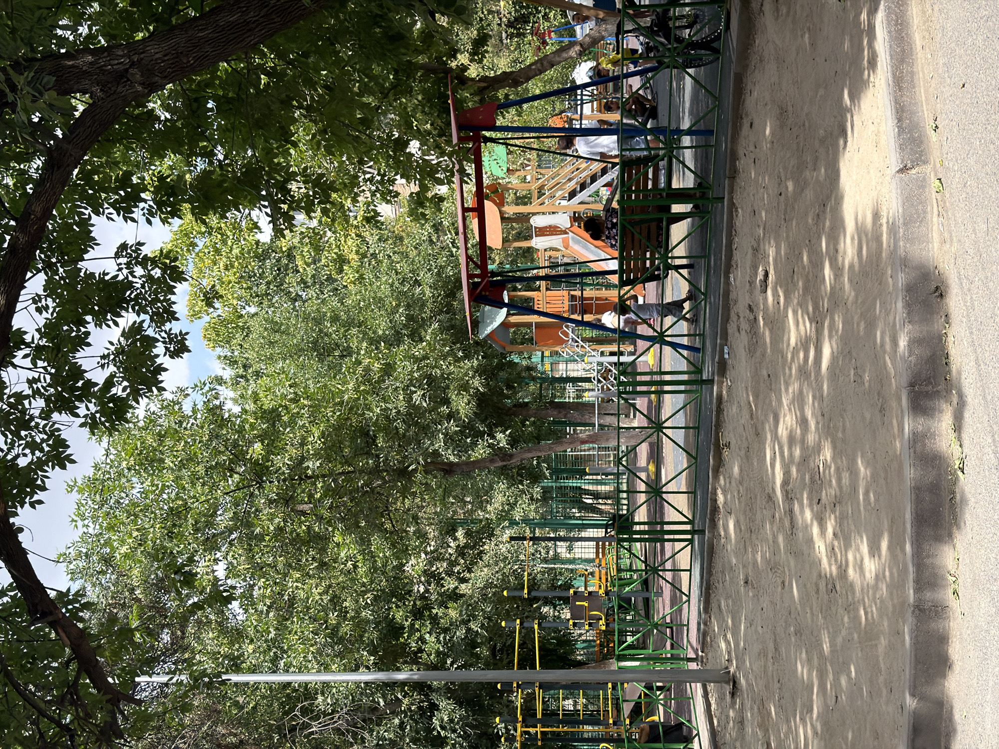 The image depicts a children's playground in Shymkent, Kazakhstan, likely a residential complex. The playground features a vibrant swing set with red and blue accents, a wooden multi-level play structure with slides and stairs, and a sandpit in the foreground. A green metal fence encloses the playground area. Several individuals, including children and adults, are visible within the playground. Some children are playing on the equipment, while others are seated or walking around. A bicycle is parked near the fence. The scene is framed by lush green trees, suggesting a warm, sunny day. The playground is situated near apartment buildings, indicated by structures visible in the background.