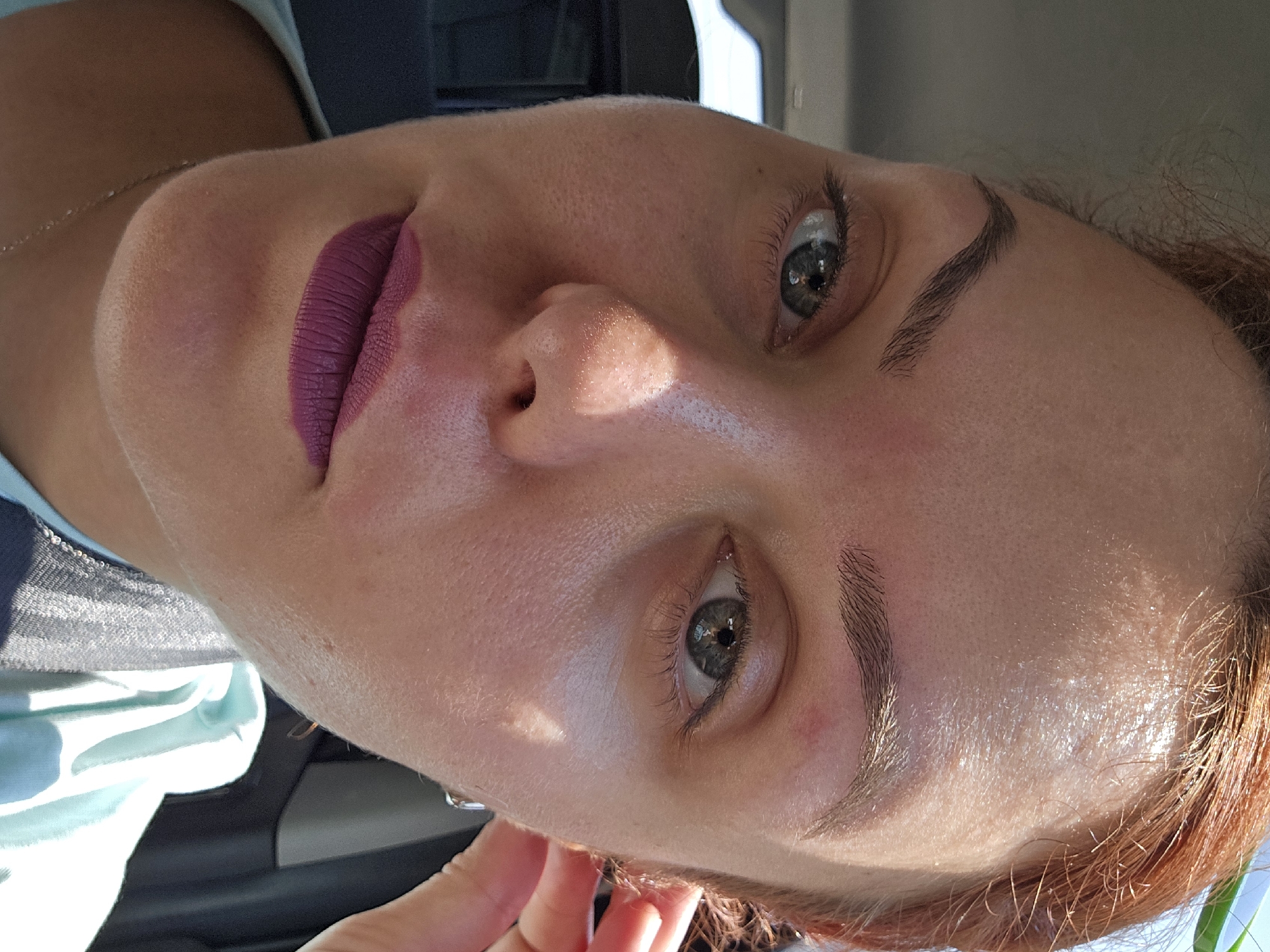 A medium close-up, front-facing, indoor shot shows a fair-skinned woman with auburn hair, wearing a light blue shirt, and purple lipstick. The woman’s right eye is partially obscured by her hand, which is touching her ear. Sunlight illuminates her face, creating a sheen on her forehead and cheeks. The background is out of focus, with hints of car interior and a bright sky. The scene is set in the city of Unknown, Unknown.