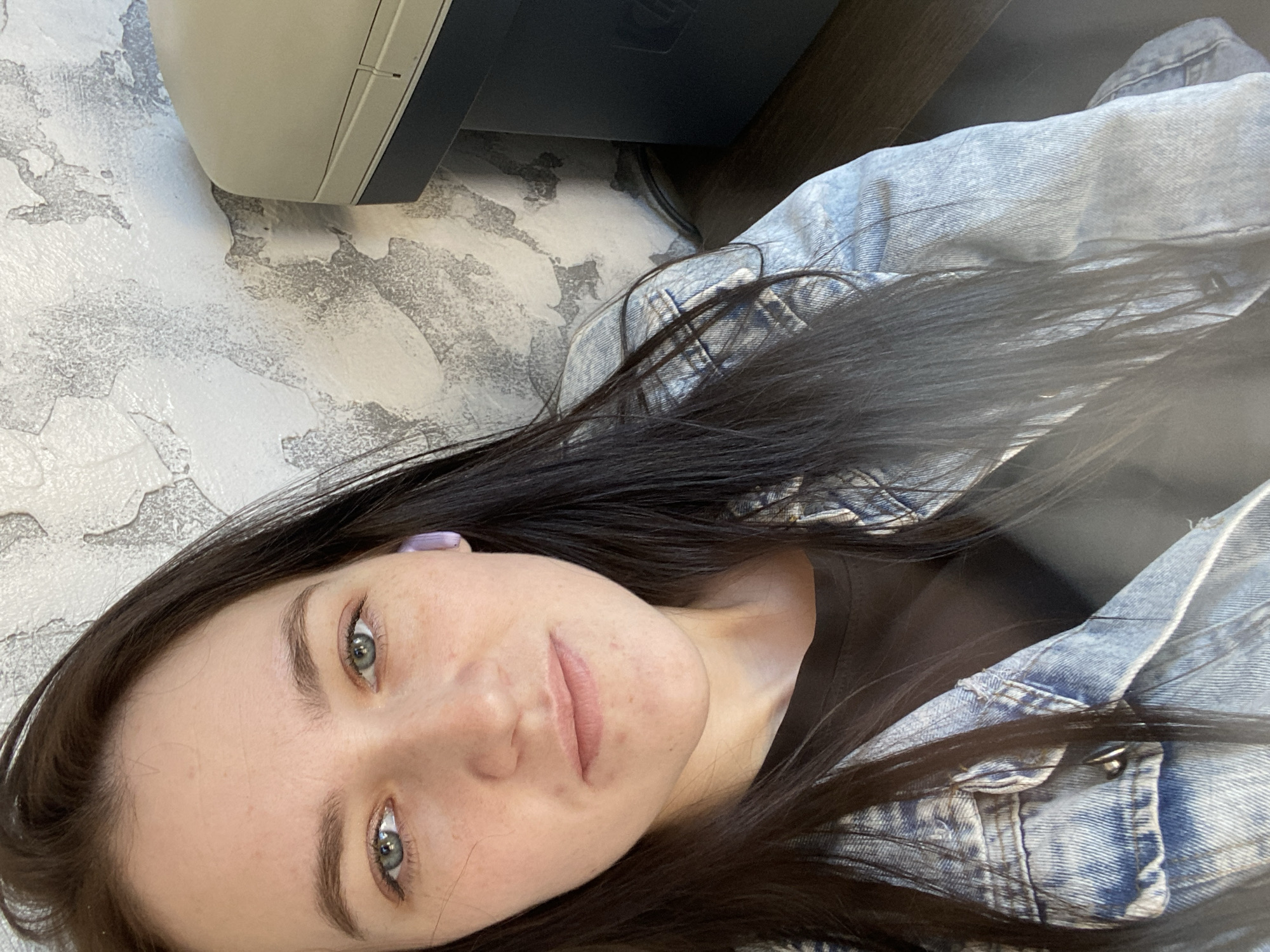 A woman with fair skin, dark hair, and light-colored eyes is posing for a selfie. She's wearing a light-wash denim jacket and a dark top. A small purple object is visible in her left ear, possibly an earbud. The background includes a light-colored textured wall and a portion of an electronic device, potentially a printer or scanner. The photograph gives no verifiable indication of the city of Chaikovsky, Russia.