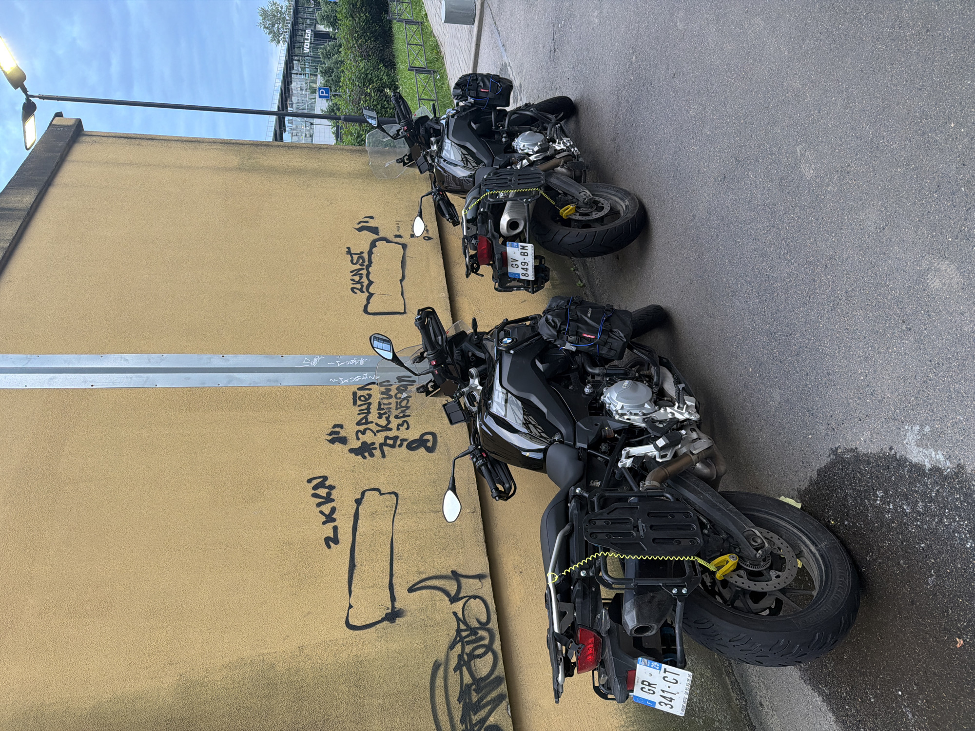 Two black BMW adventure motorcycles are parked on a wet asphalt surface next to a textured yellow wall covered in graffiti. The motorcycle in the foreground has a license plate that reads "GR 341-CT". It is equipped with panniers and a rear rack. A yellow coiled lock is attached to the rear wheel. The motorcycle in the background also has panniers and a rear rack, and its license plate is visible, reading "GV 849-BM". Graffiti on the wall includes the phrases "2KKN" and "#Зашел". A street light with two lamps is mounted on a pole behind the motorcycles, and in the background, a modern building with signage and some greenery can be seen under a cloudy sky.