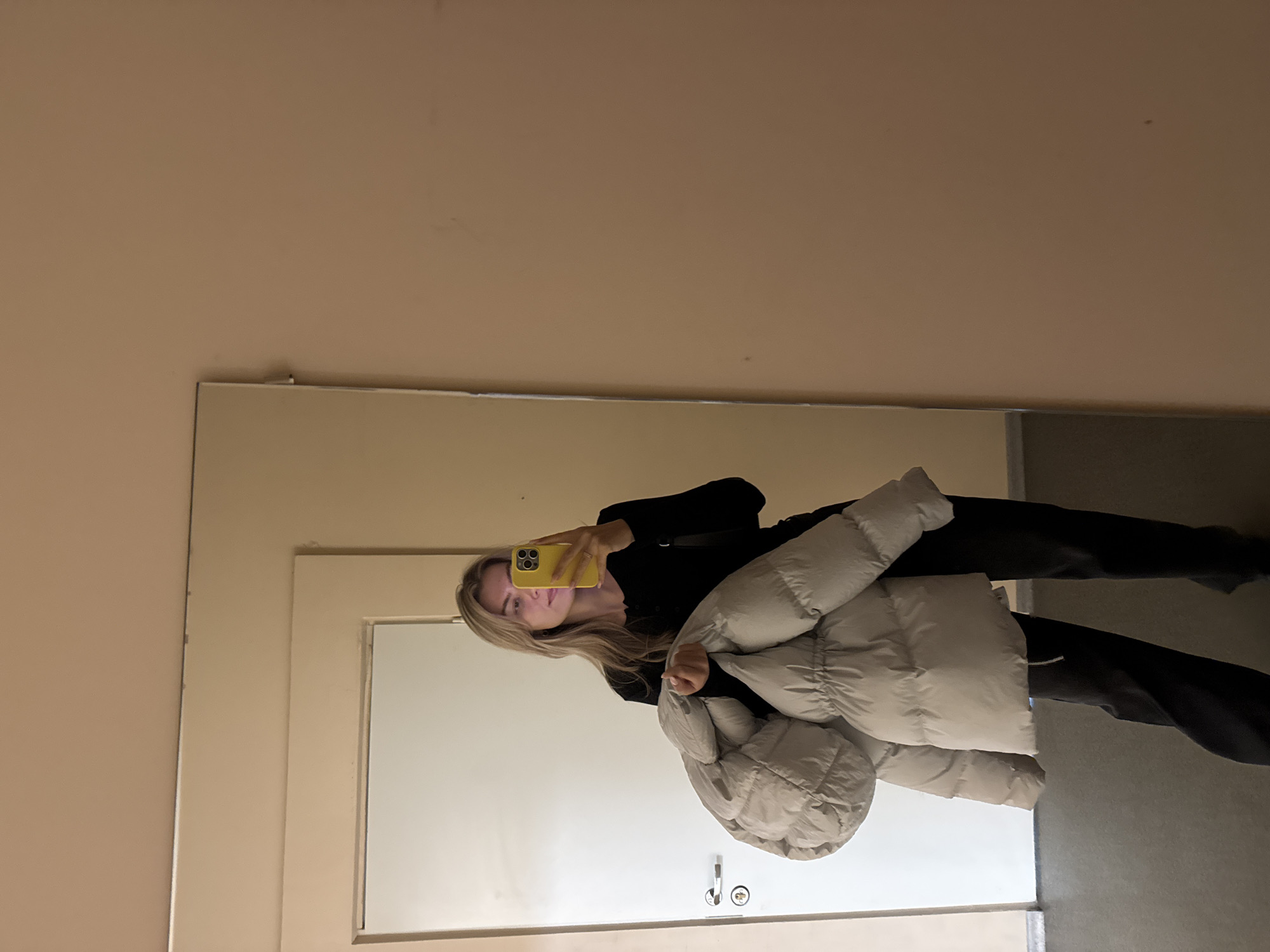 A woman is taking a selfie in a mirror. She is holding a yellow phone in her right hand. She is wearing a black long-sleeved shirt and black pants. She is holding a puffy light grey jacket in her left arm. She is standing in front of a door. The location appears to be inside a building, but the city is unknown. The walls are light tan. The floor is light grey.