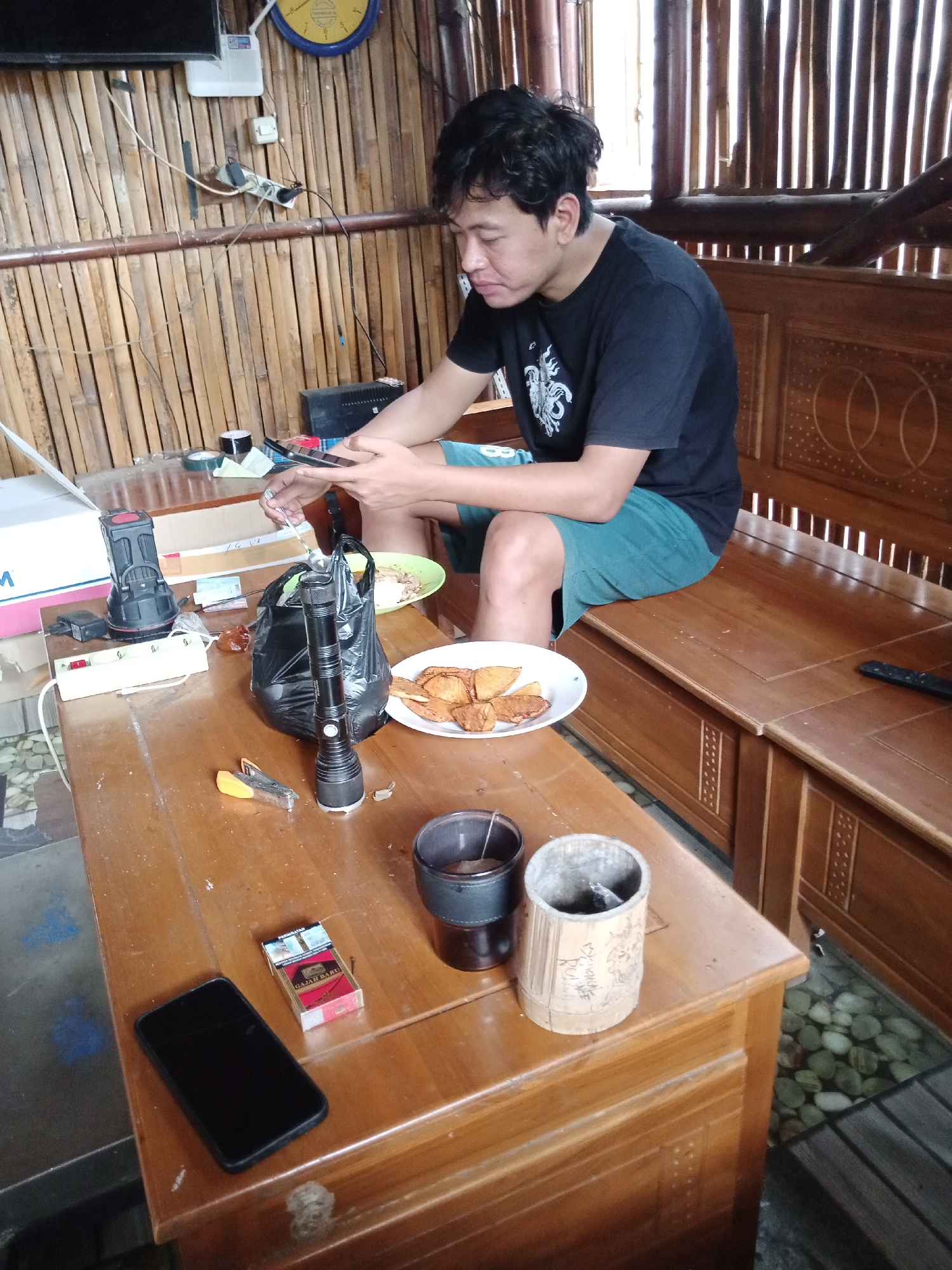 In a rustic outdoor seating area, possibly a veranda or pavilion, a young man is seated at a wooden table. The background features a wall constructed of vertical bamboo poles, with a wooden slatted roof and railings overhead, suggesting a traditional Indonesian architectural style. Sunlight filters through the gaps in the bamboo, creating a dappled effect. The man, wearing a black t-shirt and teal shorts, is engrossed in his smartphone, holding it in his left hand while his right hand is extended, holding a small object, possibly a piece of food, towards a plate of fried snacks.
On the table, various objects are scattered. To the man's left, there's a black plastic bag containing food, and a white plate with fried snacks, some of which appear to be pastels or samosas. Next to the plate are two flashlights, one black and red, and another silver one. A white power strip with multiple outlets is also visible, connected to a black adapter. In the foreground, a black smartphone lies on the table next to a pack of "GAJAH BARU" cigarettes. Two cylindrical containers, one dark and the other made of intricately carved bamboo, are also on the table. A pair of small scissors and a yellow object, possibly a stapler, are also present. The floor beneath the table is a mosaic of pebbles and dark wooden planks. The overall scene evokes a casual, relaxed atmosphere, with the man seemingly taking a break. The location is identified as Candi Wates, Indonesia.