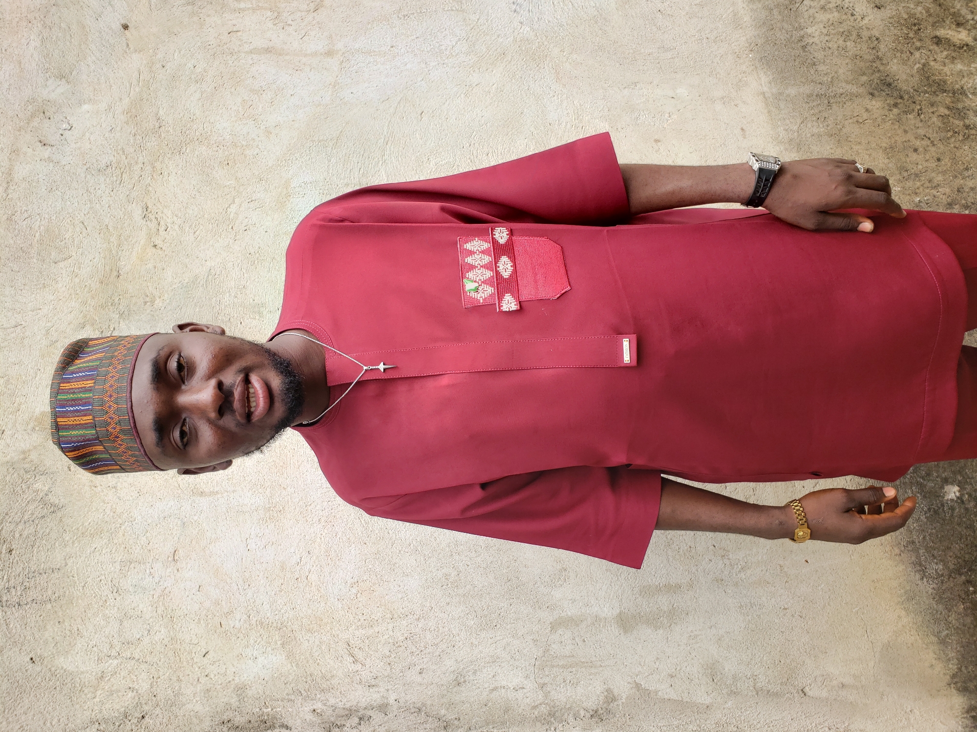 A man is standing against a textured beige wall. He is wearing a maroon, long-sleeved traditional African garment with a decorative pocket on the left side. He also wears a patterned cap on his head. A silver necklace with a pendant hangs around his neck. On his left wrist, he has a black wristwatch with embellishments, and on his right wrist, he wears a gold bracelet. He appears to be standing outdoors, though the exact location in Lokoja, Nigeria cannot be determined from the image.