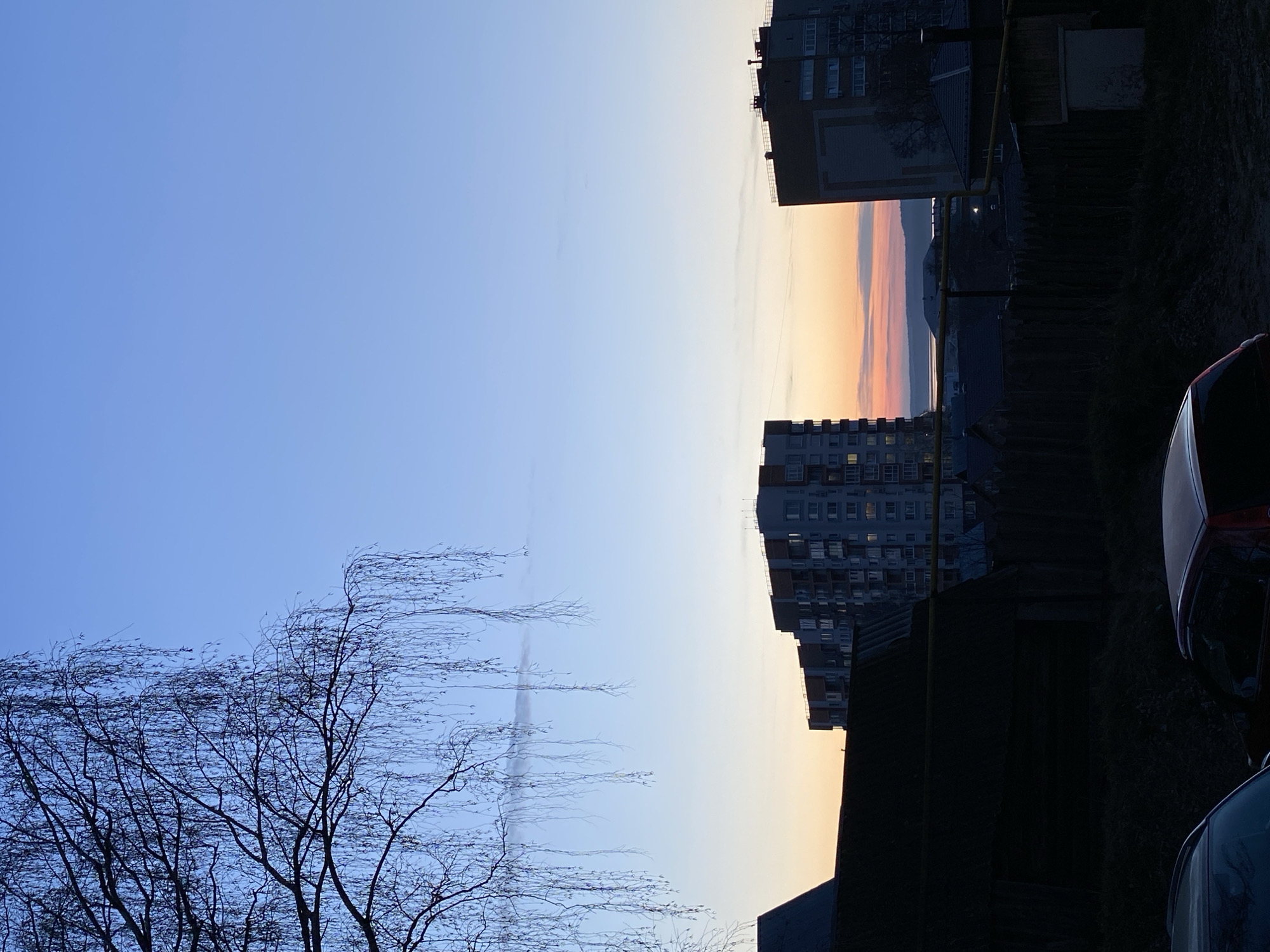 The image captures an atmospheric urban landscape in Votkinsk, Russia, at either sunrise or sunset. The sky dominates the upper two-thirds of the frame, displaying a gradient from a clear, light blue at the top to vibrant warm hues of yellow, orange, and pink along the horizon on the right. A few faint, wispy cloud formations or contrails are visible in the expanse of blue sky.
Silhouetted against this colorful horizon are several multi-story residential buildings, likely apartment blocks, which occupy the right and lower-right portions of the frame. Their facades are mostly dark due to the low light, but some windows show illuminated interiors, indicating either evening or early morning hours.
In the far left foreground, the intricate, bare branches of a deciduous tree extend upwards against the blue sky, suggesting late autumn, winter, or early spring. Further into the midground, dark, indistinct structures, possibly fences or shrubbery, contribute to the shadowed foreground. A partial view of a dark-colored car, likely red or maroon, is visible in the bottom-right corner, its roofline and rear obscured by shadow. Yellow utility pipes are also faintly visible running vertically near one of the buildings.
In the distance, just above the horizon and below the brightest part of the sky, faint silhouettes of what appear to be hills or mountains are discernible. No people or overt activities are visible, presenting a serene and quiet moment in the city. There is no visible text in the image.