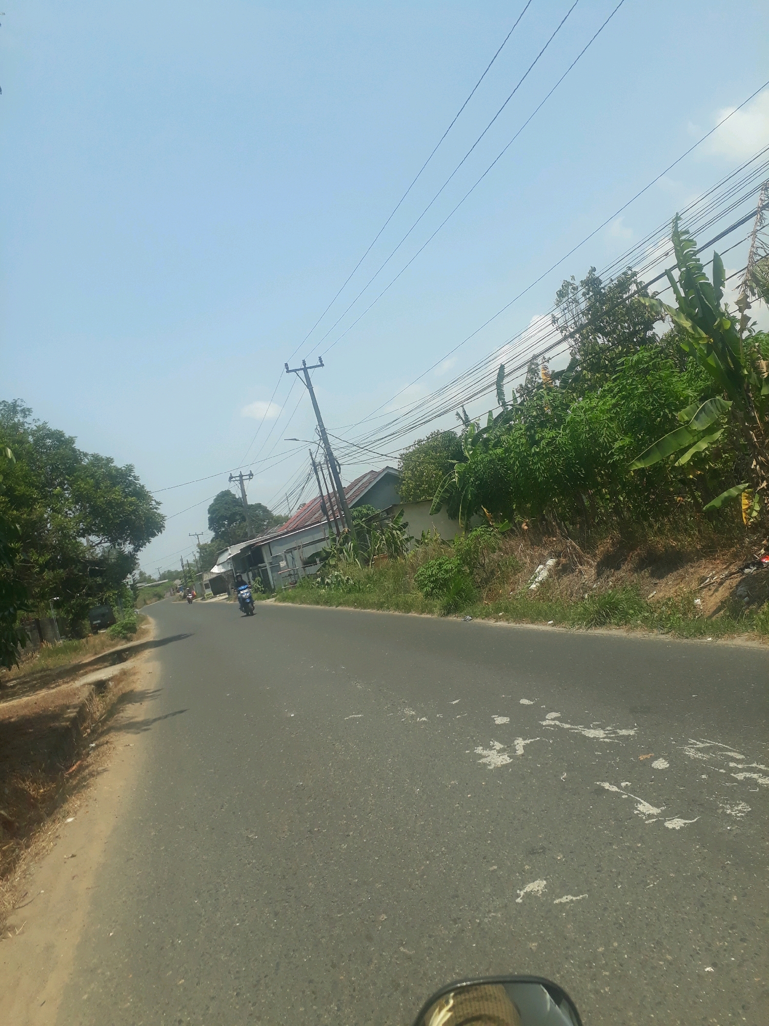 A straight, paved road curves slightly to the left in Pangkalpinang, Indonesia. The road is flanked by a sandy shoulder on the left and a grassy embankment with trees, vegetation, and houses on the right. Overhead, power lines crisscross the sky, supported by utility poles. One utility pole is prominently visible on the right side of the road. A blue and white scooter is traveling on the road in the middle ground, heading away from the viewer. Several other vehicles and structures are visible further down the road. The sky is clear and blue, with a few wispy clouds. The scene appears to be a typical street in a tropical or subtropical urban area.
