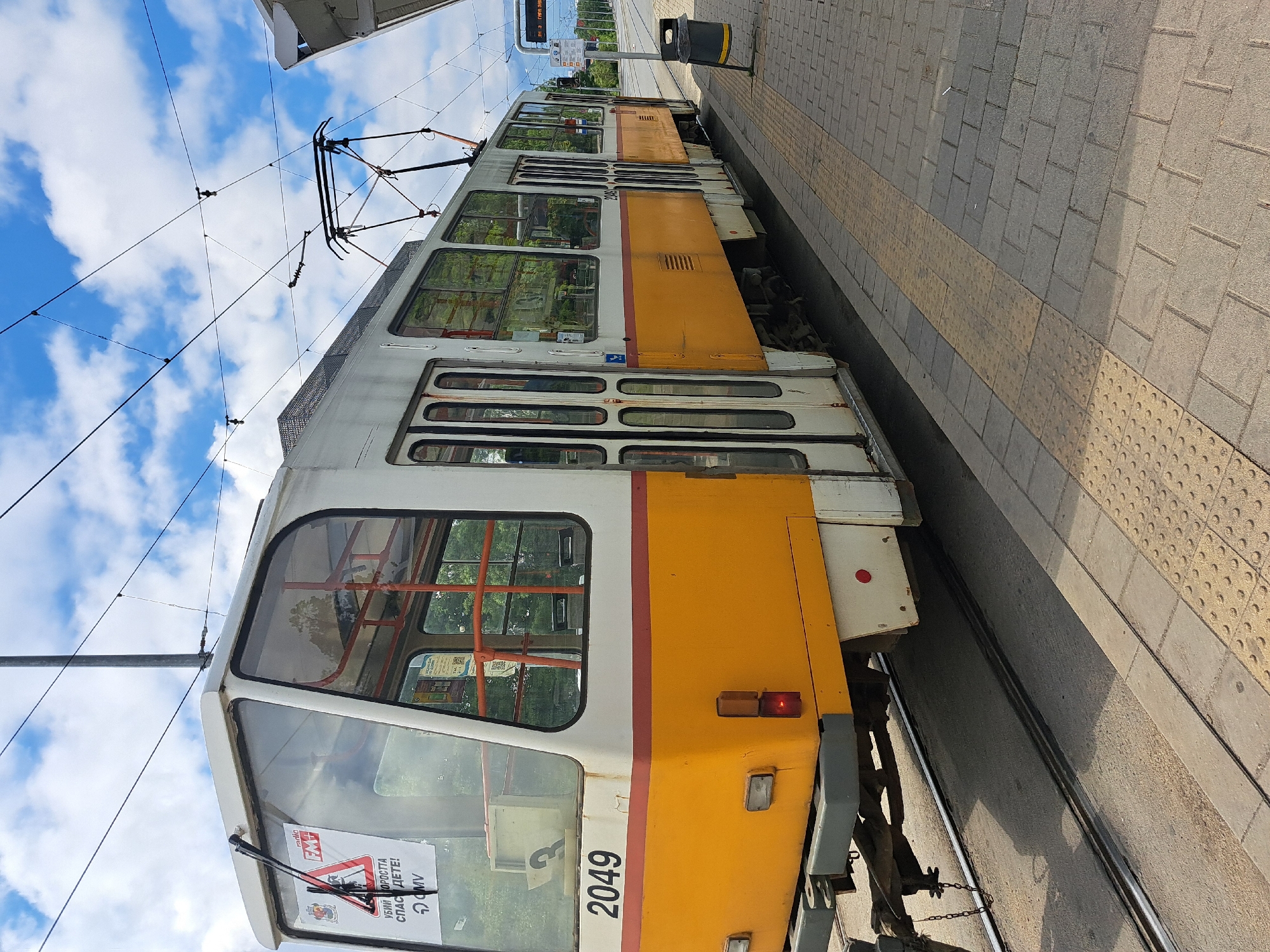 The image shows a partially visible yellow and white tram, number 2049, stationary at a tram stop. The tram is facing right and is positioned beside a gray brick platform with a yellow tactile paving strip along the edge. Overhead, there are tram wires and supports against a backdrop of blue sky with scattered white clouds. A trash can is on the platform behind the tram. The presence of Cyrillic text on a warning sticker on the tram window, as well as the presence of typical trams from the Sofia network suggests the location is Sofia, Bulgaria.