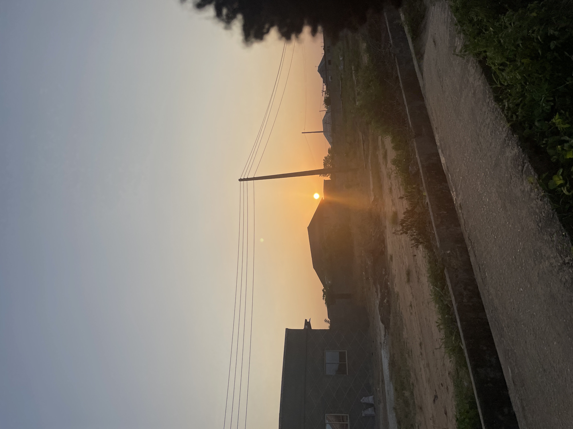 The image captures a serene sunset scene in Iyafin, Nigeria. A vast, clear sky transitions from a pale blue at the top to a warm orange hue near the horizon, where the sun is setting. In the mid-ground, several low-rise buildings with sloped roofs are visible, appearing to be under construction or recently built. One building on the left has a grey, textured exterior and several windows. A prominent wooden utility pole stands to the right of center, with multiple power lines stretching horizontally across the frame. The ground is a mix of dirt and sparse vegetation, with a concrete structure running along the bottom of the frame, possibly a drainage ditch or a pathway. In the foreground, a paved surface is visible, with green foliage growing along its edge. The overall atmosphere is peaceful and quiet, indicative of the end of the day in a developing area. No people are present in the image.