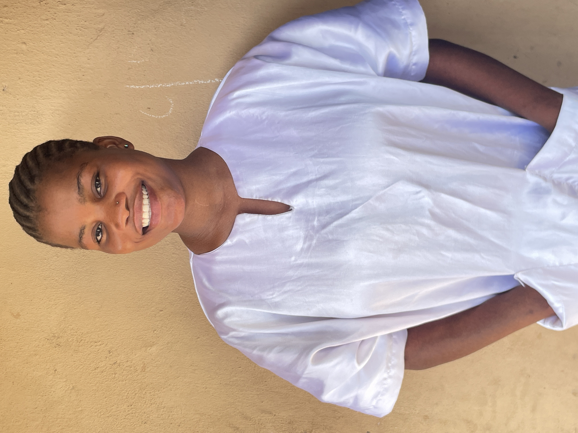 The image, taken in Badagry, Nigeria, features a young woman smiling brightly and looking directly at the camera. She has dark skin and her hair is styled in neat, intricate braids. A small silver nose ring adorns her left nostril, and a subtle earring is visible in her right ear. She is dressed in a loose-fitting, lustrous white garment, possibly a robe or traditional attire, which reflects light, creating soft highlights that suggest a silky or satin-like texture. Her arms are visible, partially covered by the flowing sleeves of her clothing.
The background consists of a textured, light brown or tan wall, indicating an outdoor or semi-outdoor setting. Faint, irregular white chalk markings are visible on the wall to the left and above her shoulder, but they do not form discernible words or clear images. The lighting suggests it is daytime, casting no harsh shadows, implying a bright but diffused light source. The woman's wide, cheerful smile conveys a clear sense of happiness and warmth.