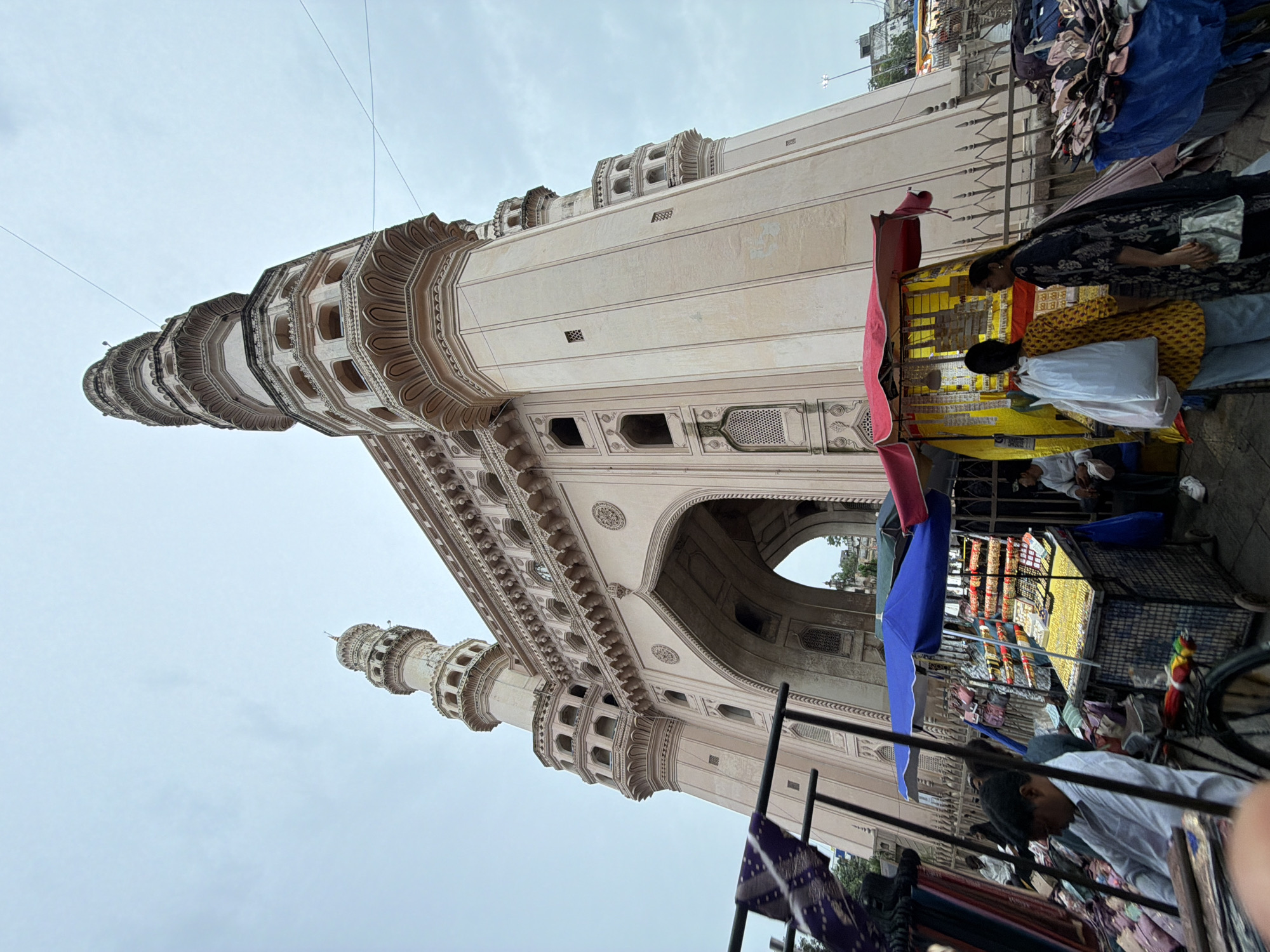 The image shows a low-angle view of the Charminar in Hyderabad, India, against an overcast sky. The iconic monument, a square building with four arched entrances and four minarets, is rendered in a light-colored stone. In the foreground, several market stalls are set up, displaying various goods, including textiles and accessories. People are seen browsing and interacting with vendors. Some are seated, while others are standing. A cart filled with fabrics is also visible, suggesting a bustling street market environment. The overall scene depicts a lively urban setting characterized by historical architecture and commercial activity.