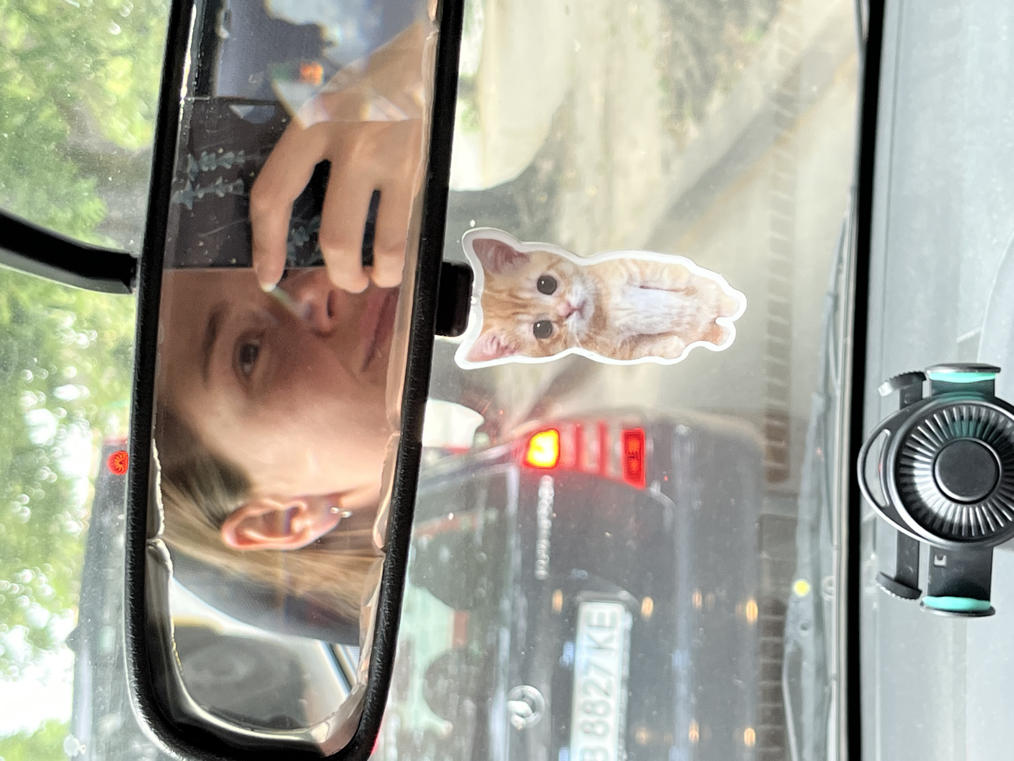 A selfie is taken in the rearview mirror of a car, showing a person holding a phone and a fluffy orange kitten keychain hanging from the mirror. The car is in traffic in Orizari, Bulgaria, evidenced by the license plate "B 8827 KE." The image is a close-up, with the driver's face and the kitten keychain as the primary subjects. The background shows other cars and trees, suggesting an outdoor setting.