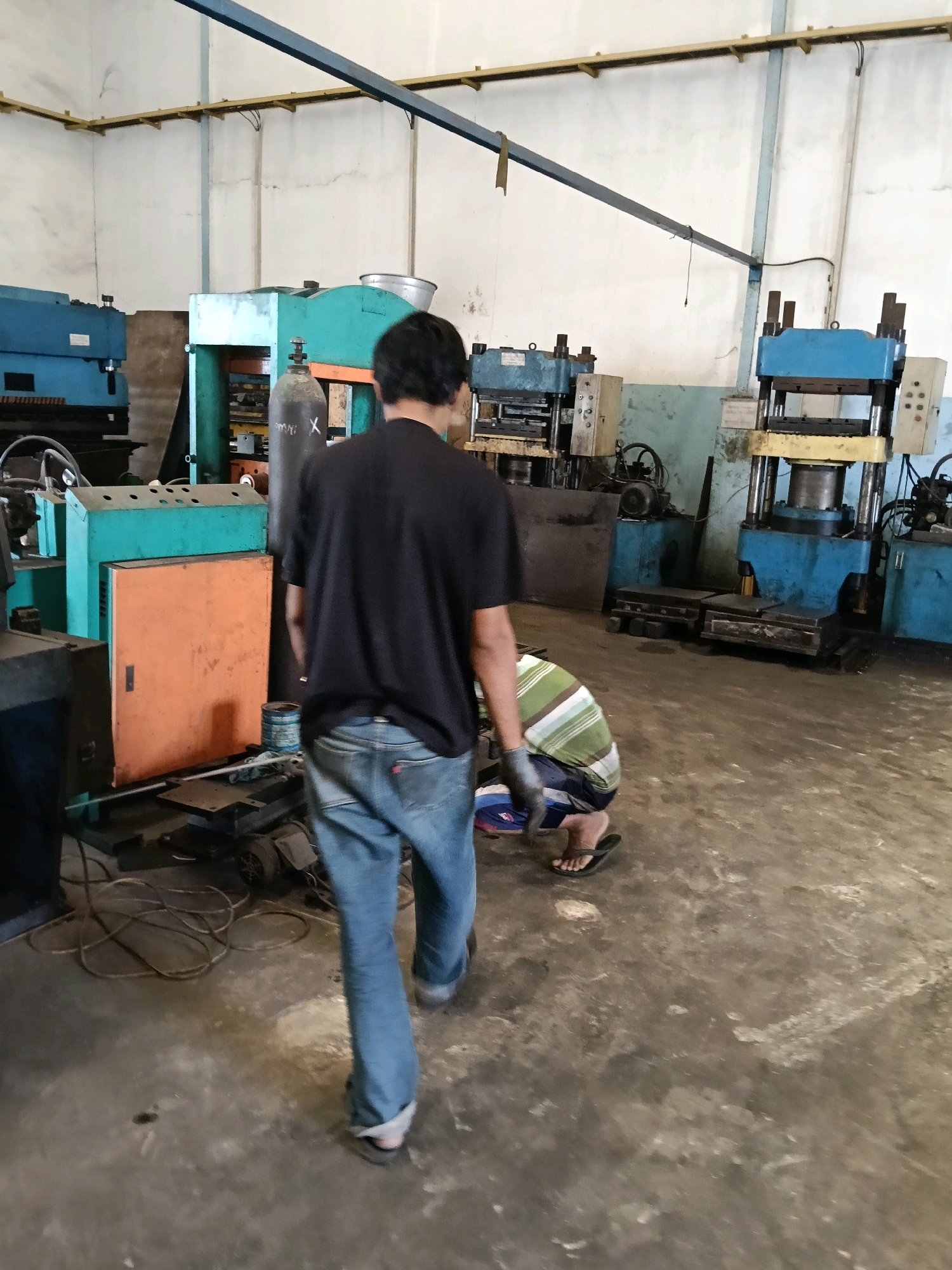 The image depicts an indoor industrial setting, possibly a workshop or factory, located in Belimbing, Indonesia. The floor is concrete and shows signs of wear and dirt. Various pieces of heavy machinery are visible, including what appear to be hydraulic presses and other industrial equipment, some painted blue and teal.
In the foreground, a person wearing a black t-shirt and blue jeans is walking away from the camera. Their posture suggests they are moving through the space. Further into the scene, another person is crouched down, seemingly working on something on the floor. This individual is wearing a horizontally striped t-shirt in green and white, dark shorts, and flip-flops. They have black gloves on their hands.
Other objects present include a gas cylinder, electrical wiring, coiled cables, and a toolbox or storage unit. The walls appear to be painted a light color, and a blue metal beam runs horizontally across the upper part of the image, supported by yellow brackets. The overall impression is of an active, functional industrial environment.