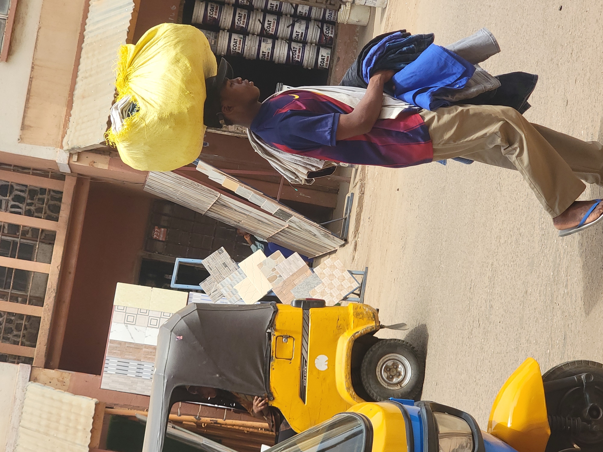 A man in a burgundy and blue striped shirt and tan pants balances a large yellow sack on his head and carries a stack of clothing in his left arm. He is walking on a paved street next to a yellow three-wheeled vehicle (tuk-tuk). Behind him, stacked tiles are visible, and in the background, there are rows of stacked cans labeled "NECOAT," an indication he is near or in a merchant district. The building behind him has a sign with the number 737 on it. The scene likely takes place in Maiburiji, Nigeria.
