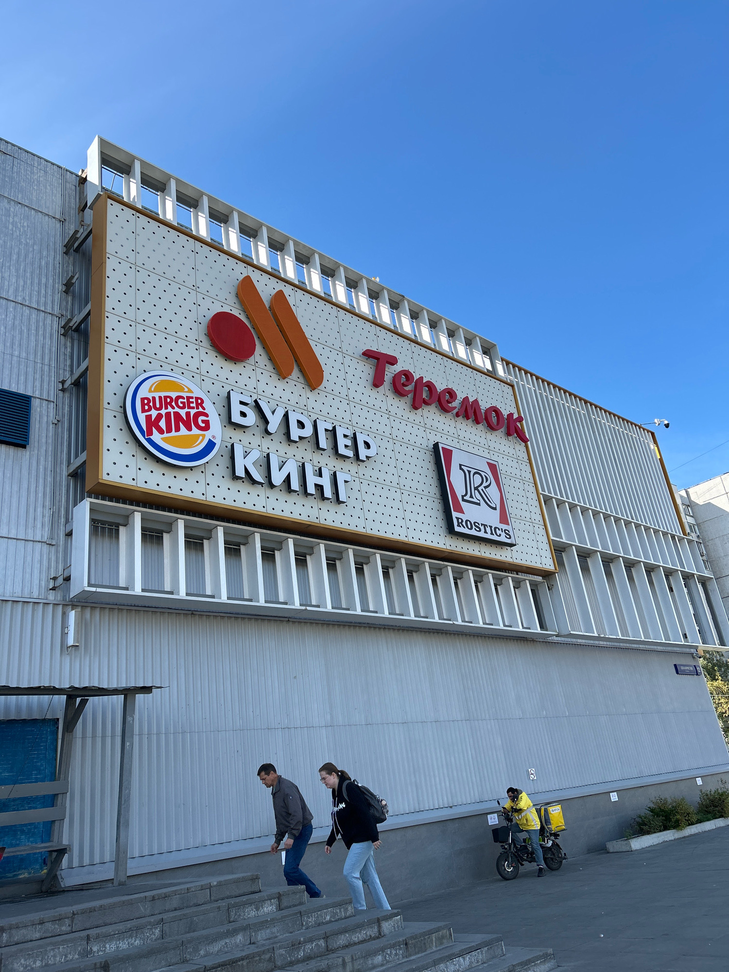 In Moscow, Russia, a bright blue sky forms the backdrop for a multi-story building featuring prominent signage for fast-food establishments. A large white facade is adorned with the Burger King logo on the left, accompanied by Russian text reading "БУРГЕР КИНГ". To the right, the "Теремок" logo, featuring stylized orange shapes and a red circle, is visible, along with the "Rostic's" logo. The building's architecture includes vertical white louvers along its upper sections and a corrugated metal siding on the lower portions. In the foreground, concrete stairs lead up to the building. Three individuals are present: a man and a woman walk up the stairs, and a person in a yellow jacket on a scooter with a yellow delivery bag is parked on the sidewalk to the right, looking down at their phone.