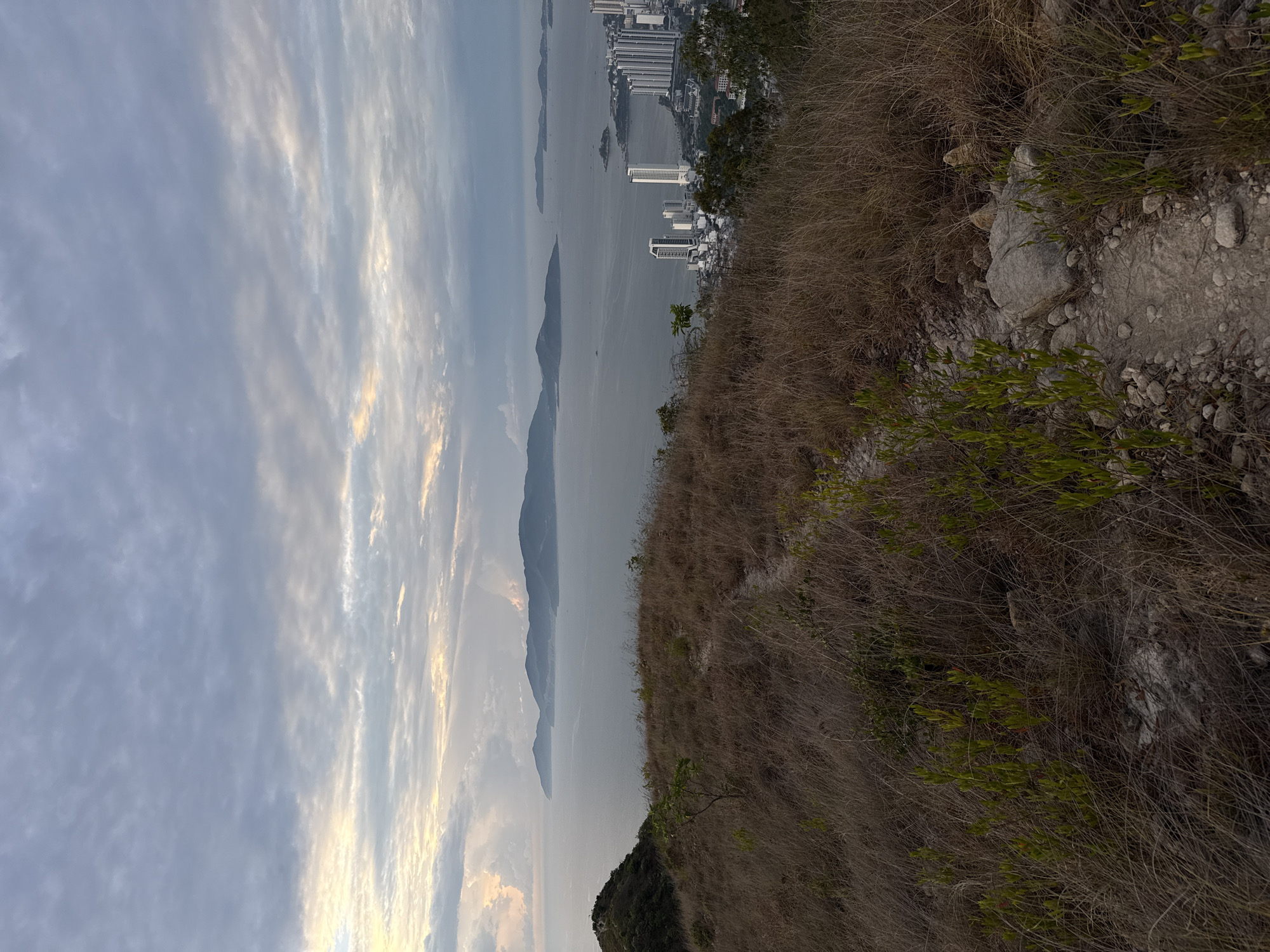 The image presents a landscape view taken from an elevated position, possibly a hillside. To the left, the sky dominates with cloud formations, some tinged with sunlight. A body of water extends into the distance, containing several islands or landmasses. On the right side of the image, vegetation is prominent, consisting of dry grass, rocks, and some green foliage. In the distance, a portion of a city skyline is visible, featuring high-rise buildings. The environment appears to be a natural setting near an urban area. The scene suggests a viewpoint from a natural, elevated position looking towards a coastal cityscape. The location is Nha Trang Ward, Vietnam.
