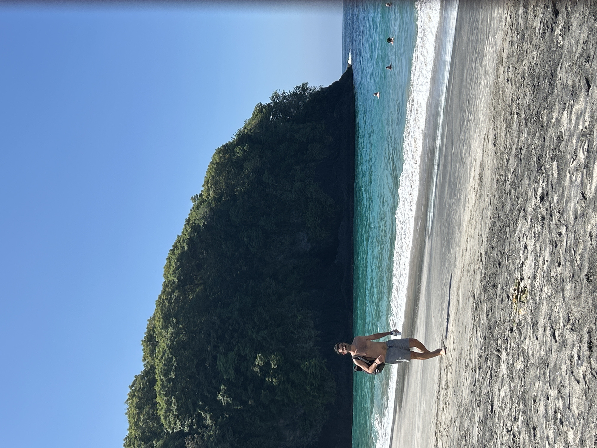A lone man walks on a sandy beach in Karangasem, Indonesia, with a green, tree-covered cliff rising behind him. The ocean is a vibrant turquoise, with small waves lapping the shore and a few people swimming in the distance. The man, wearing grey shorts and carrying a black backpack, appears to be walking towards the viewer. The sky is a clear, bright blue. The sand is dark and speckled with grey and white.