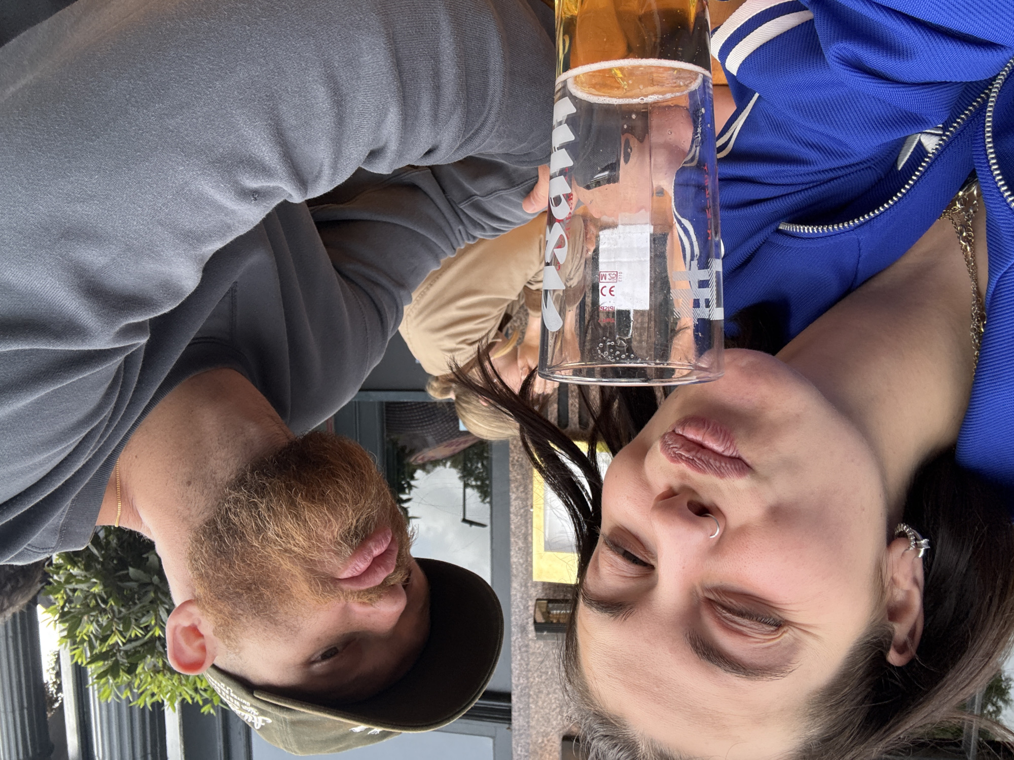 The image captures a close-up selfie of two young adults, a female and a male, seated outdoors at a table, likely at a pub or restaurant. The female, positioned on the left, has dark hair, fair skin, and wears a blue track jacket, a gold chain necklace, a small patterned hoop earring in her left ear, and a silver hoop nose ring. Her eyes are closed in a squint, and her lips are pursed in a "duck face" expression. She holds a clear pint glass containing a golden-amber liquid, consistent with lager or beer, topped with a foamy head. The glass is branded with "Heineken" text and logo.
The male, positioned on the right, has fair skin, a full ginger beard, and wears a dark green baseball cap and a grey long-sleeved sweatshirt, along with a gold chain necklace. His lips are also pursed forward, mimicking the female's expression. A third individual with light-colored hair and a tan jacket is partially visible in the background, seated behind the main subjects.
The environment includes a dark-colored building facade with architectural details, a partially visible yellow sign or board, and green foliage. The sky is overcast. The outdoor setting and architectural elements are consistent with an urban environment in the United Kingdom, specifically compatible with a pub or eatery within the city of London, although no unique identifying landmarks for a specific London location are present.