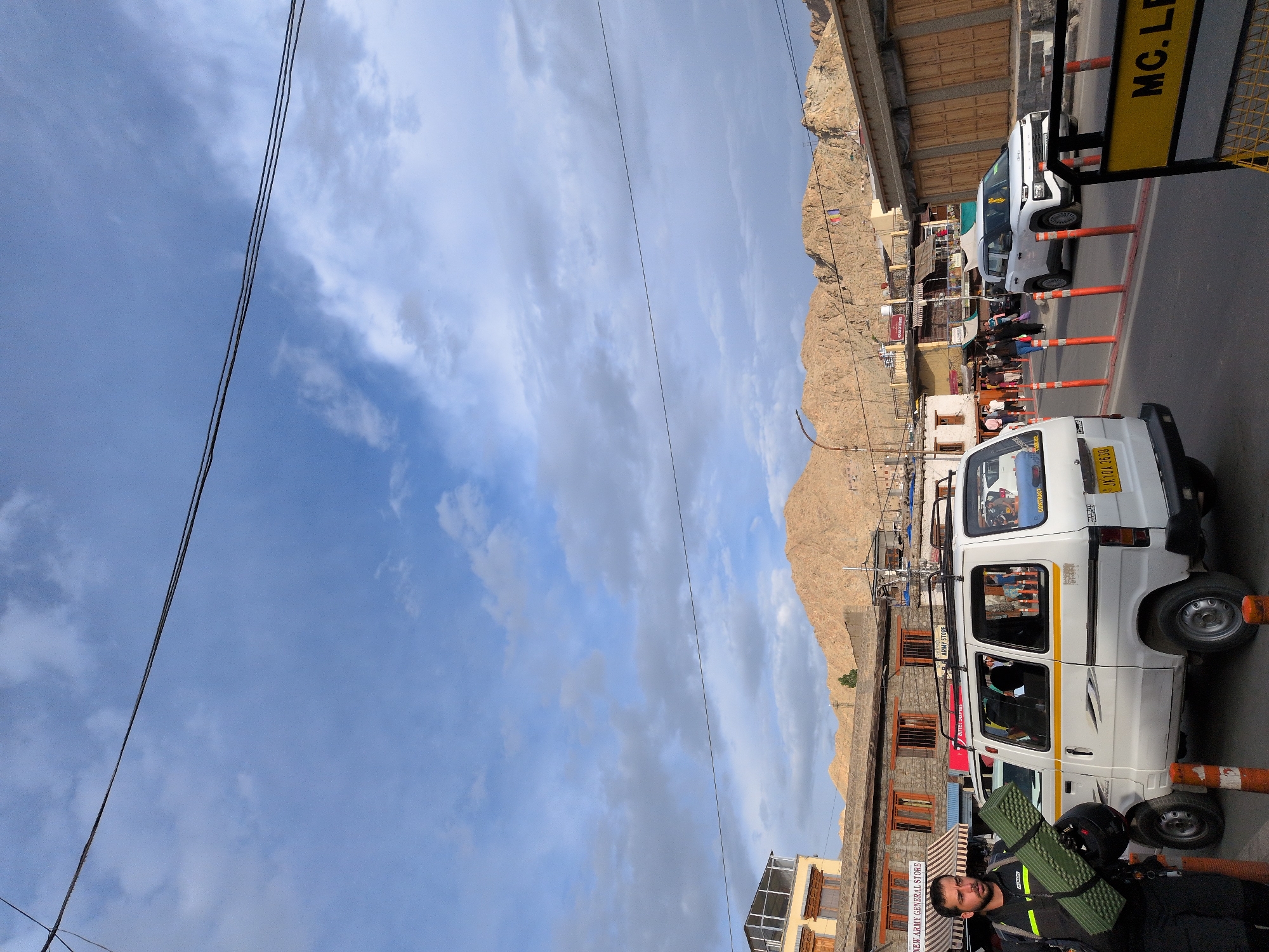 The image captures a street scene in Leh, India, under a cloudy sky. In the foreground, a man walks on the left side of the frame, carrying a rolled-up green sleeping mat and a black bag. To his right, a white van is parked. The van has a yellow stripe along its side and a visible license plate that reads "JK10A 3530". Orange traffic cones are placed on the road.
Further down the street, a white SUV is visible. The street is lined with buildings, some of which appear to be shops. In the background, rugged, tan-colored mountains dominate the landscape. The architecture of the buildings suggests a mountainous region, consistent with the location of Leh. A yellow sign with black lettering, partially visible on the right side of the image, reads "MC. LE". The overall scene depicts a busy street in a town nestled among mountains.
