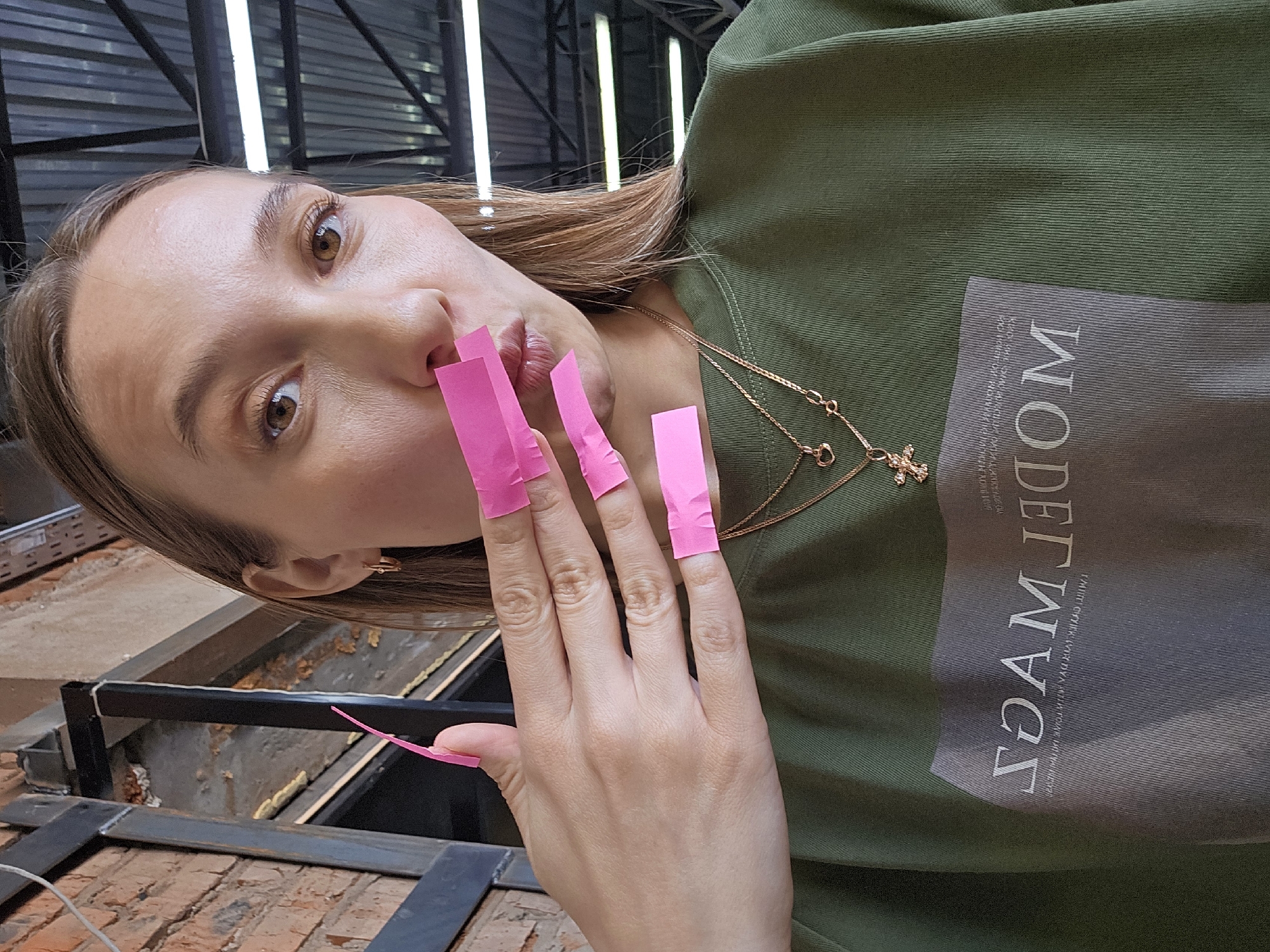 A young woman poses for a selfie in what appears to be an industrial or commercial setting, possibly in Chaikovsky, Russia, although this cannot be confirmed from the image alone. She is wearing a green shirt with a gray patch on which the reversed text "MODERN" is printed. She is also wearing a gold necklace with a cross pendant and small hoop earrings. Her fingernails have bright pink strips of tape attached. The background includes exposed brick, metal beams, and fluorescent lighting, suggesting a building under construction or renovation.