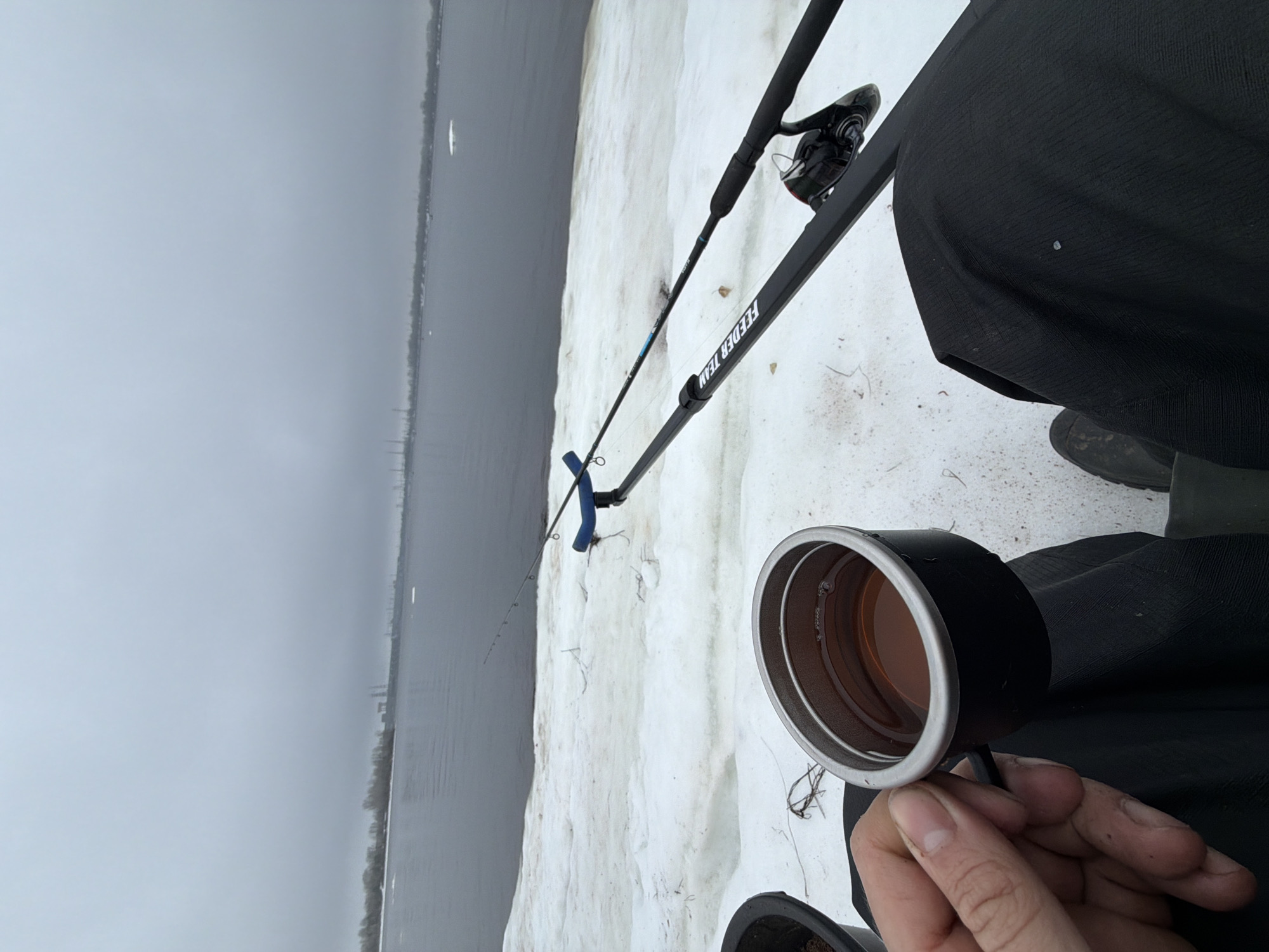A first-person perspective shot captures a person ice fishing. The scene includes a snow-covered area leading to a body of water, possibly a frozen lake or river, under an overcast sky. A fishing rod, bearing the inscription "FEEDER TEAM," is positioned near the edge of the ice, with the fishing line extending into the water. The person holding the camera is also holding a mug filled with a brownish liquid, presumably a hot beverage. The person is wearing dark clothing, with a glimpse of dark-colored footwear. The distant skyline suggests a city or industrial area along the shoreline. Based on visual cues and provided location context, this is likely a scene of ice fishing near Noviy, Russia.
