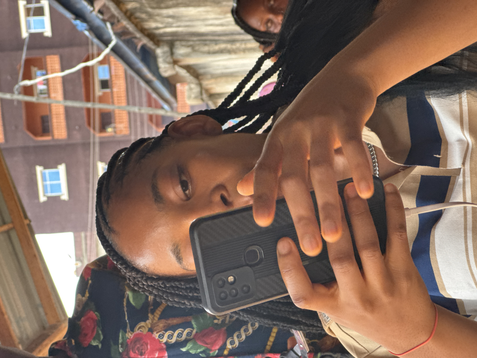 A young Black woman with braided hair is holding a black smartphone in front of her face. She is wearing a striped shirt and a silver chain necklace. Her fingers are visible around the phone. Another person is partially visible behind her. The background shows a building with orange brick and white-framed windows, typical architecture in Onitsha, Nigeria. The scene appears to be taken outdoors during daylight hours.