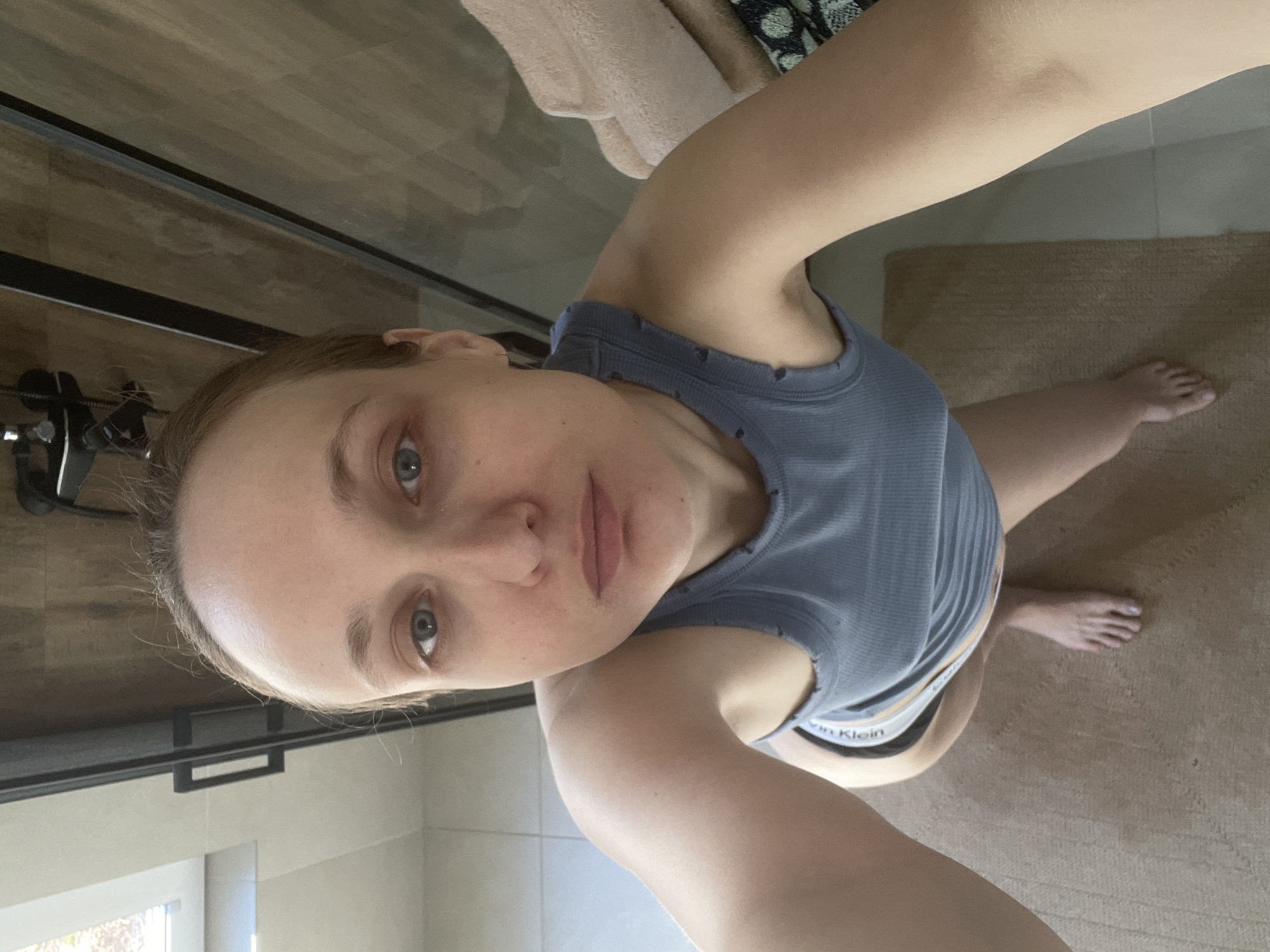 A woman is taking a selfie in a bathroom. She is wearing a gray tank top and black Calvin Klein underwear is visible. The bathroom features a glass shower door, light-colored tile walls, a beige bathmat, and wooden-look flooring. A window is visible in the background. The location is unspecified and cannot be determined from the image. The city of Подшивалово, Russia, cannot be confirmed.
