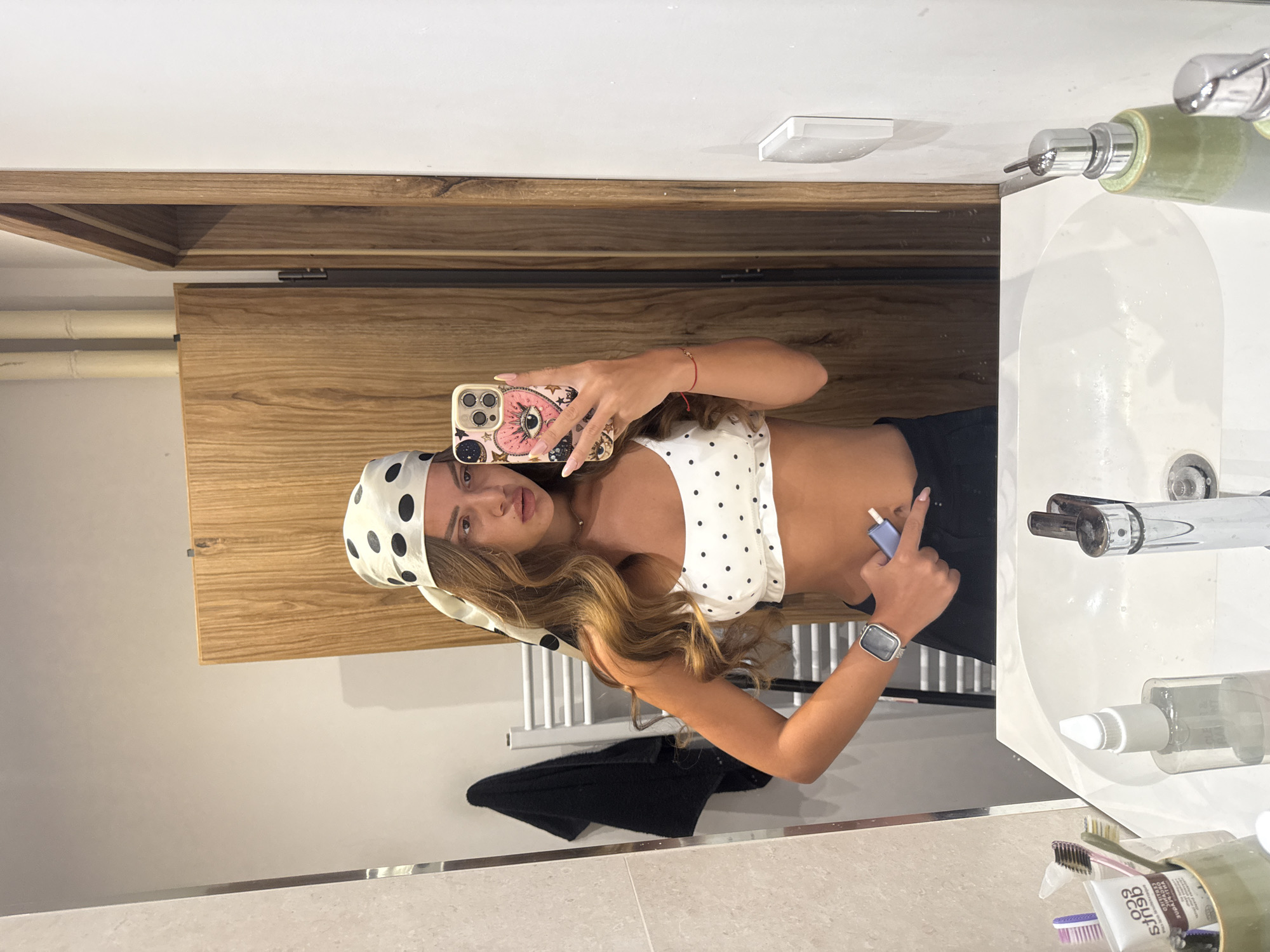 A young woman with long, wavy brown hair, wearing a white and black polka dot bandana and a matching polka dot crop top, stands in a bathroom and takes a selfie in a mirror. She is holding a phone with a heart-shaped case featuring an eye design and a blue object in her other hand. She has a smartwatch on her left wrist. The bathroom appears modern, with wooden cabinets, a white sink, and various toiletries visible. The scene is indoors, in what could be a residential bathroom. There is no indication of a specific city or location, therefore no mention of Plovdiv, Bulgaria.